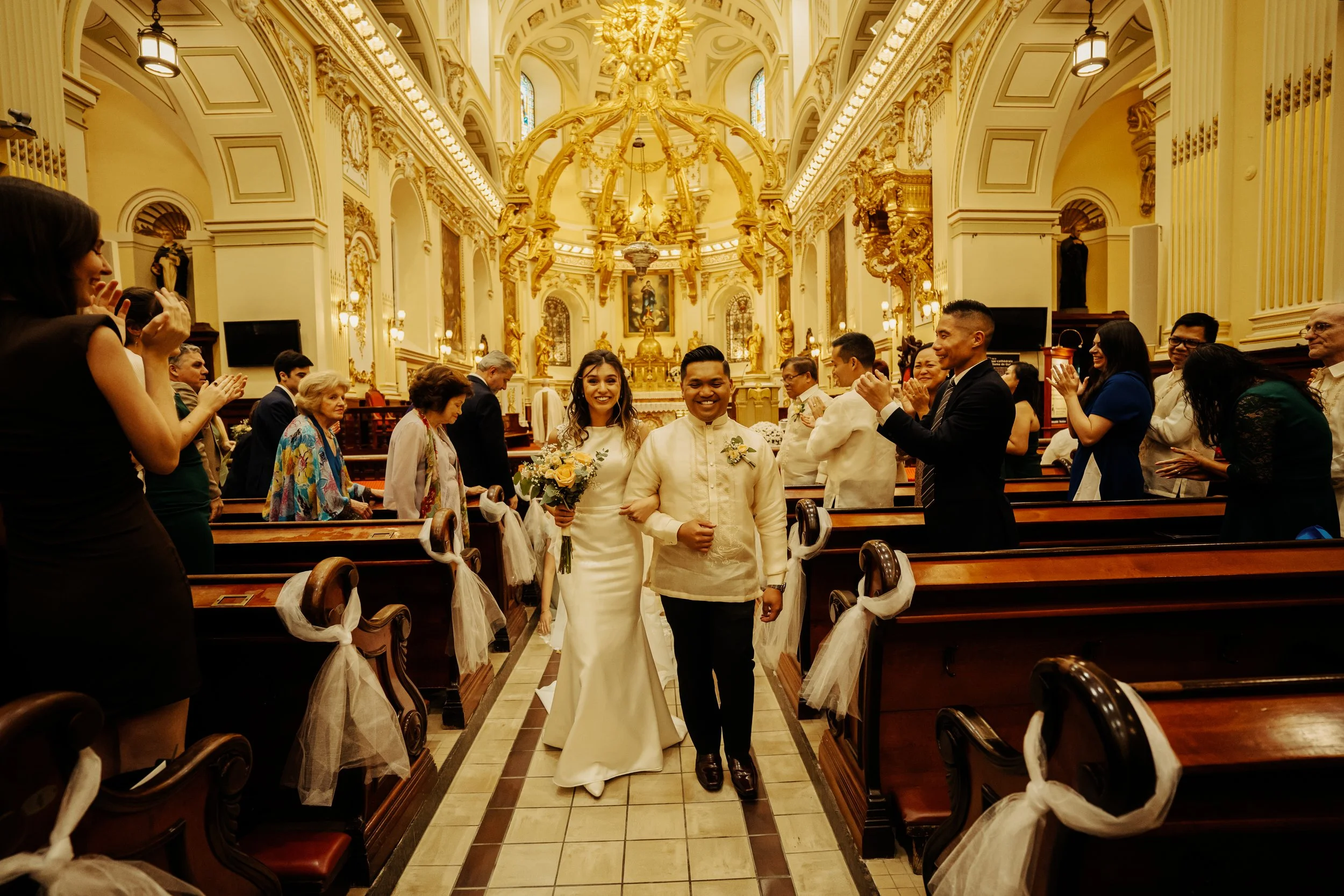 Bride and groom walking down the aisle in a church, surrounded by clapping guests, with ornate gold decorations and high ceilings in the background.