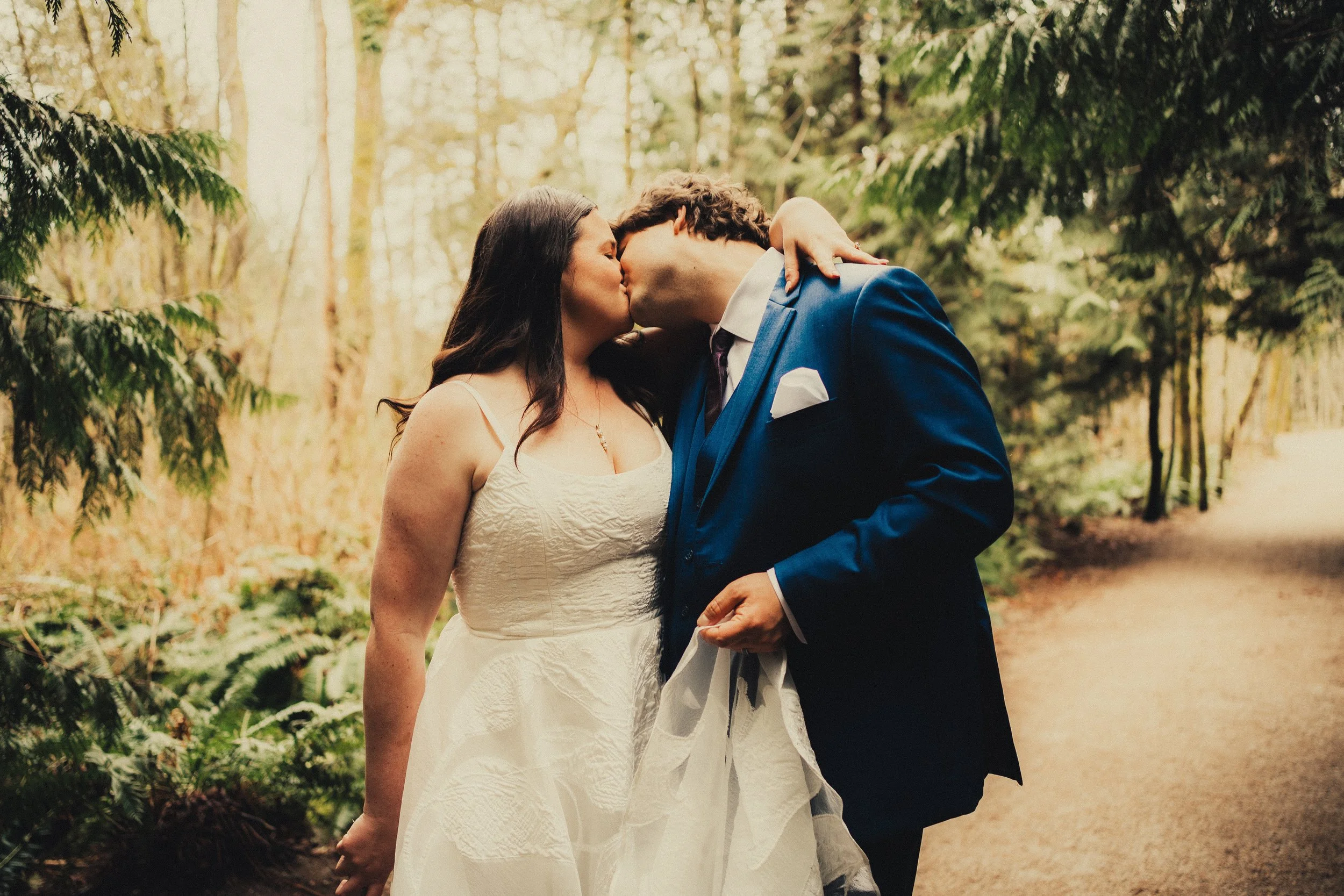 A bride and groom sharing a kiss in a forest during their wedding, with the bride in a white dress and the groom in a blue suit.