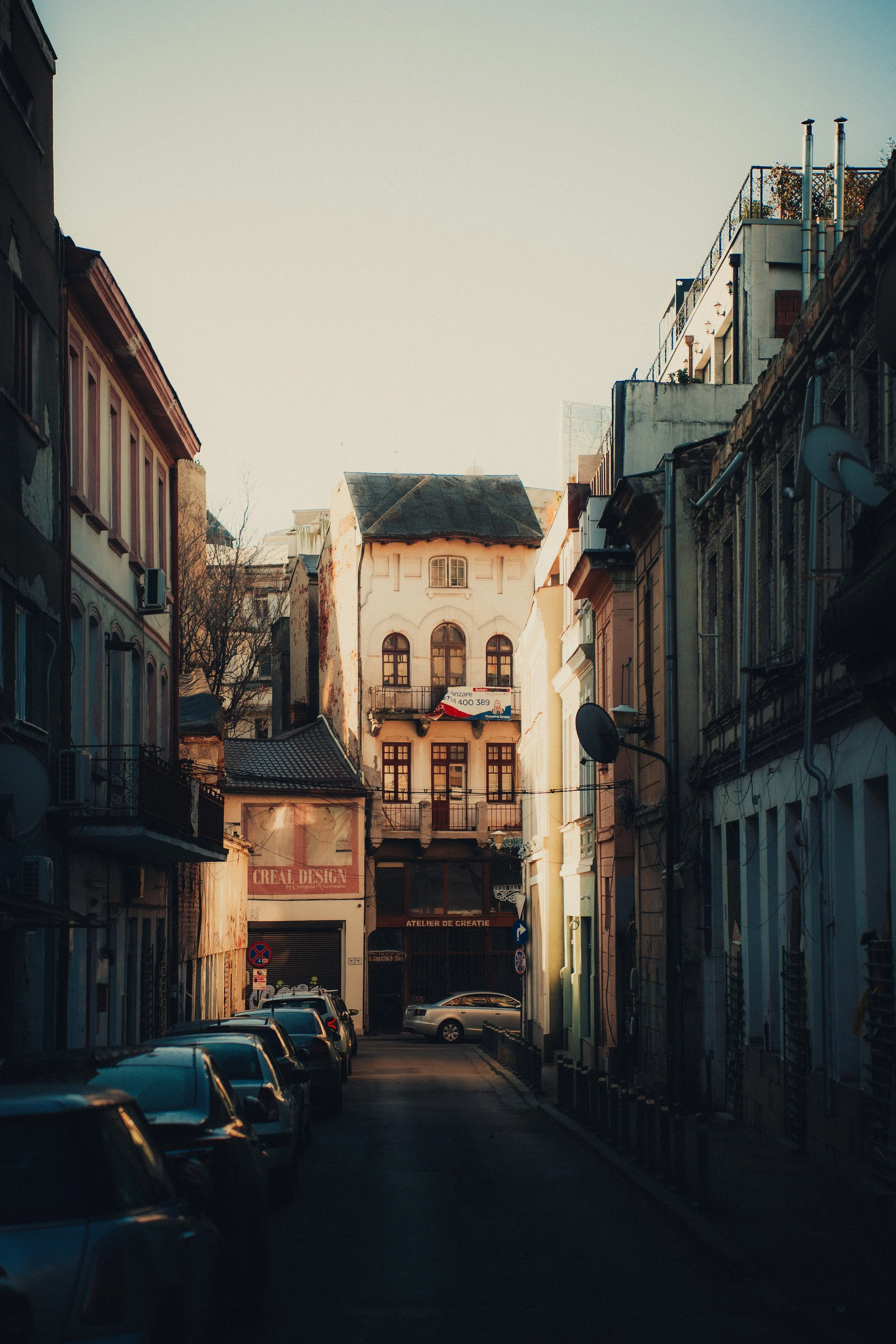 A narrow urban street lined with parked cars and old buildings, leading to a small building with a balcony and a sign that reads 'ATELIER DE CREATION.' The scene is lit by warm, late afternoon sunlight.