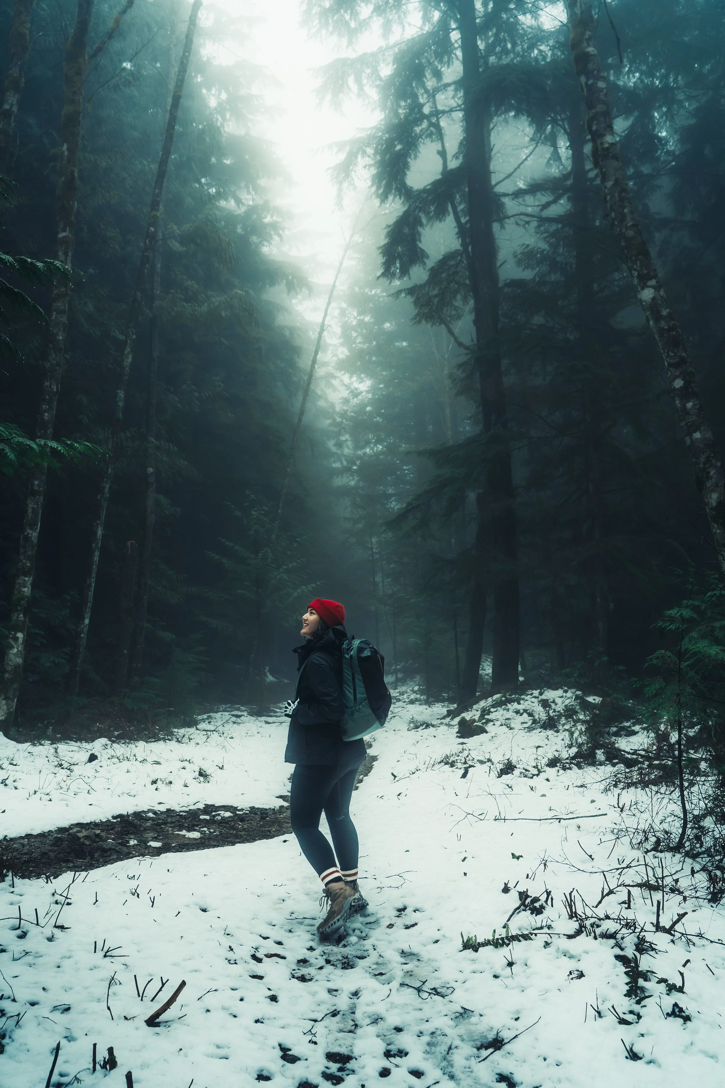 A woman hiking through a snowy forest with tall trees and foggy atmosphere, wearing a red beanie, a backpack, and winter hiking gear.
