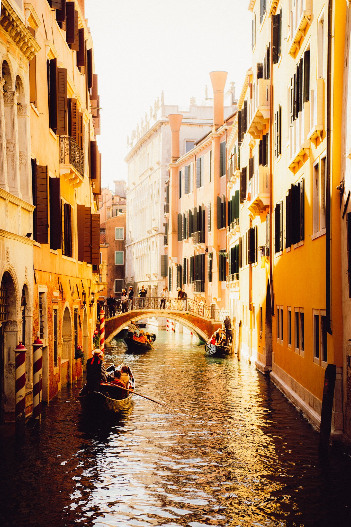 Gondolas on a canal with colorful Venice buildings on either side and a small stone bridge crossing overhead, during golden hour sunlight.