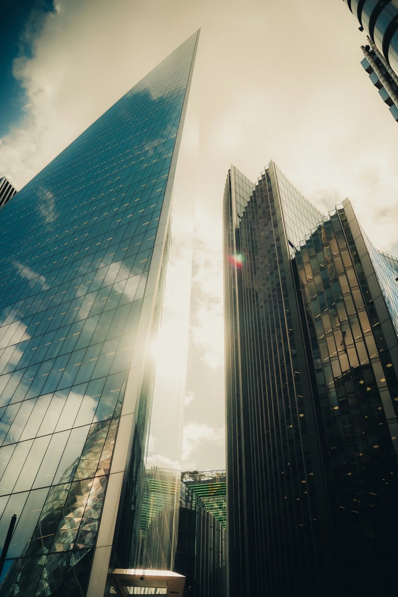 Tall modern glass skyscrapers in London reflecting the sky and clouds, with sunlight shining through.
