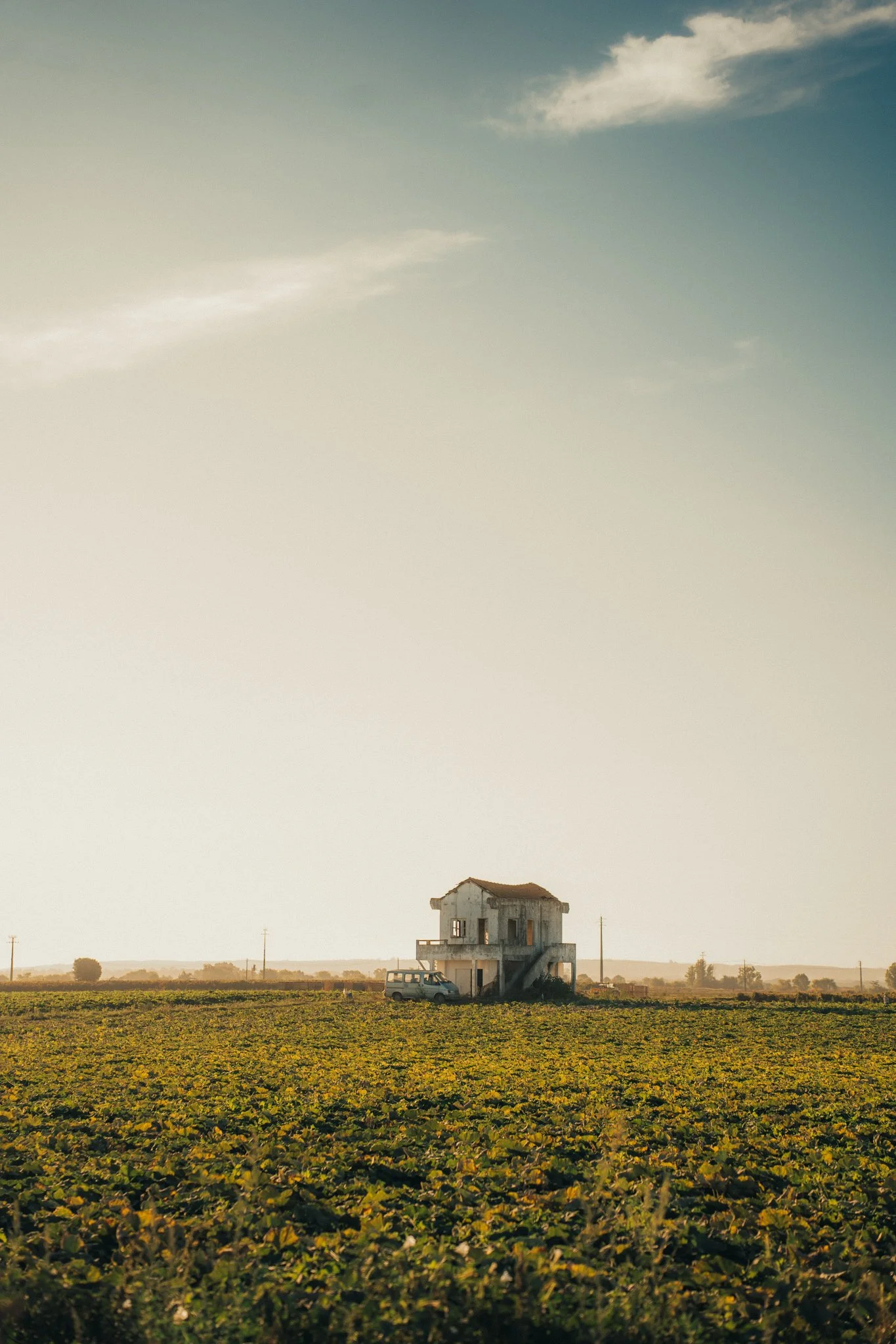 A two-story, weathered house with a garage on the ground floor, located in the middle of a large farm field with a vintage van parked nearby. The sky is partly cloudy with a mix of sun and shadow.
