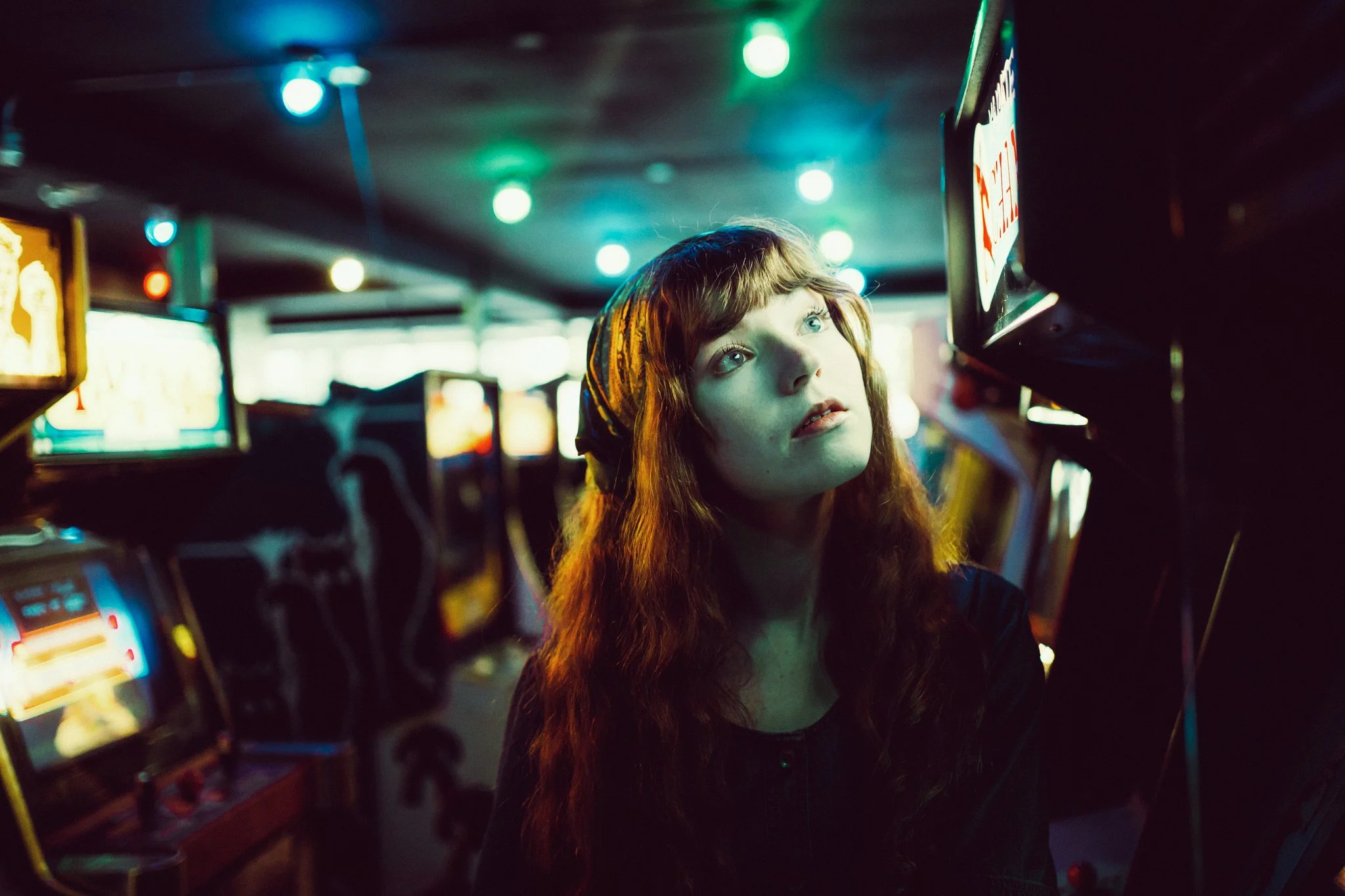 A young woman with long red hair and a headscarf looks up thoughtfully in a neon-lit arcade filled with video game machines.