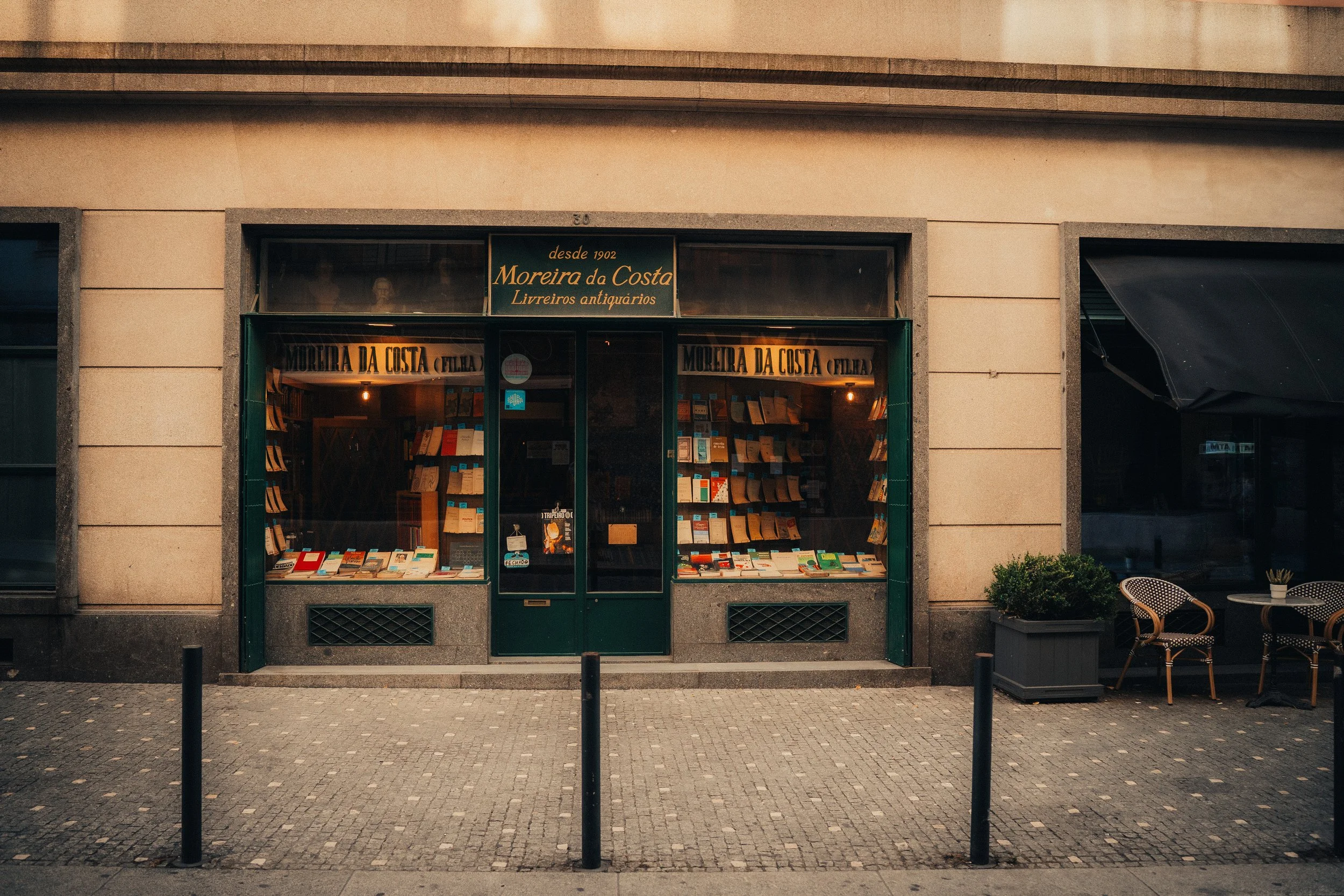 A bookstore named Moreira da Costa with books displayed in the window, located on a city street with outdoor seating and planters.