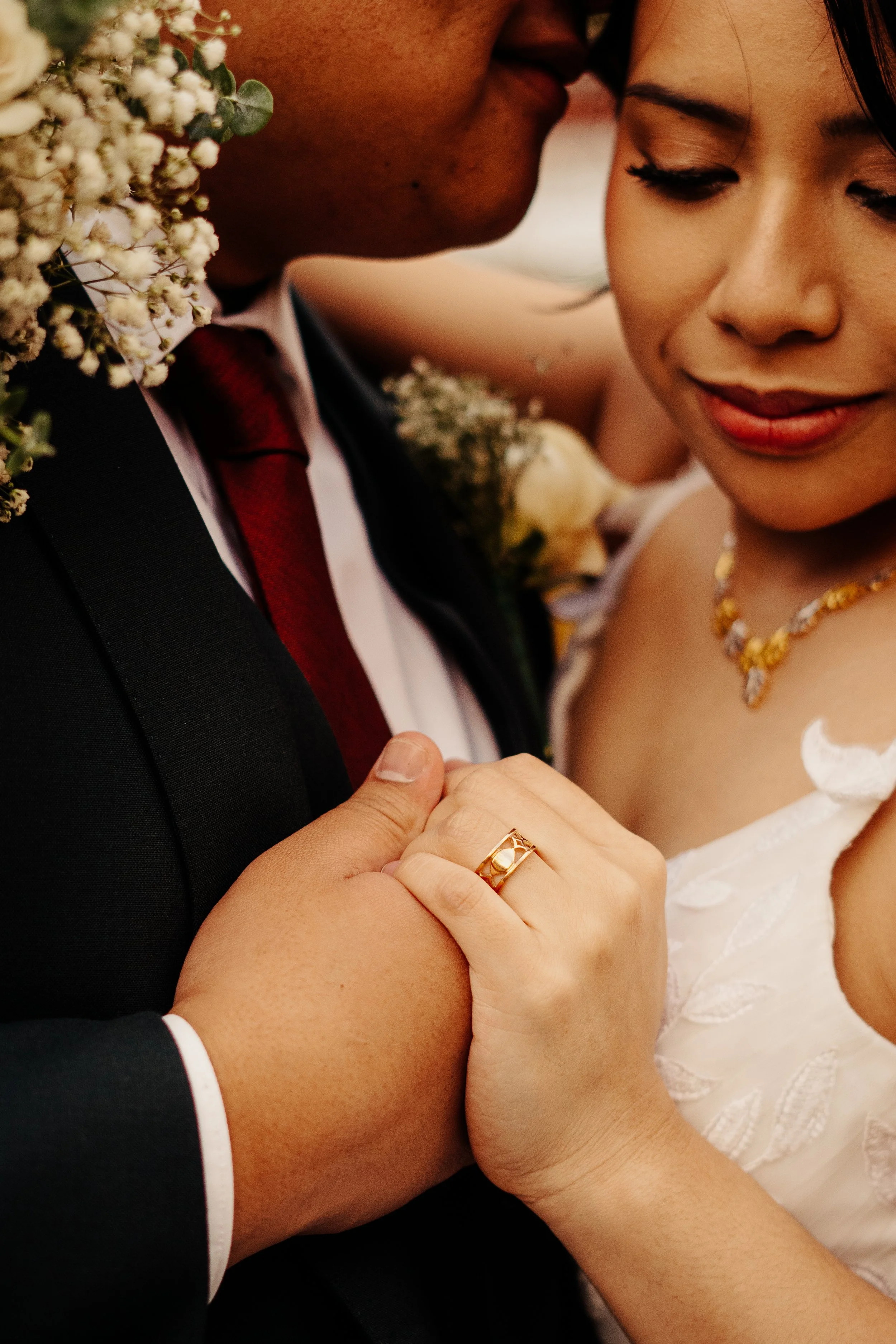 Close-up of a couple embracing at their wedding, showing part of a man's face and suit, and a woman's face with wedding dress, gold jewelry, and wedding ring. The woman is gently holding the man's arm.
