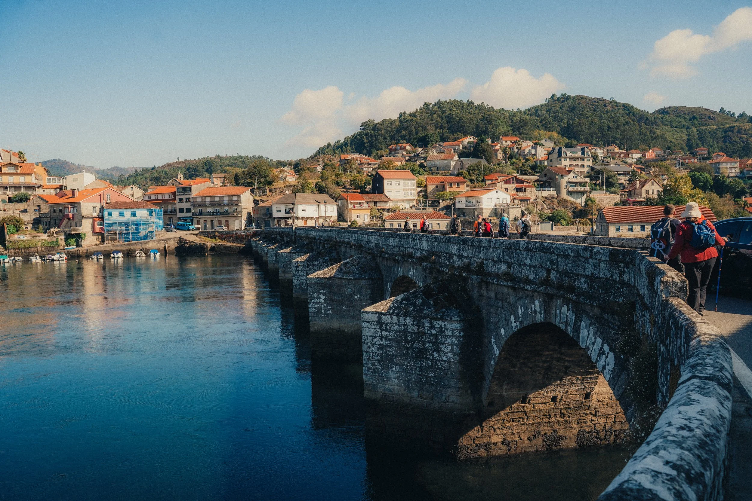 Stone bridge crossing a river with houses and hills in the background, people walking along the bridge, blue sky with some clouds.