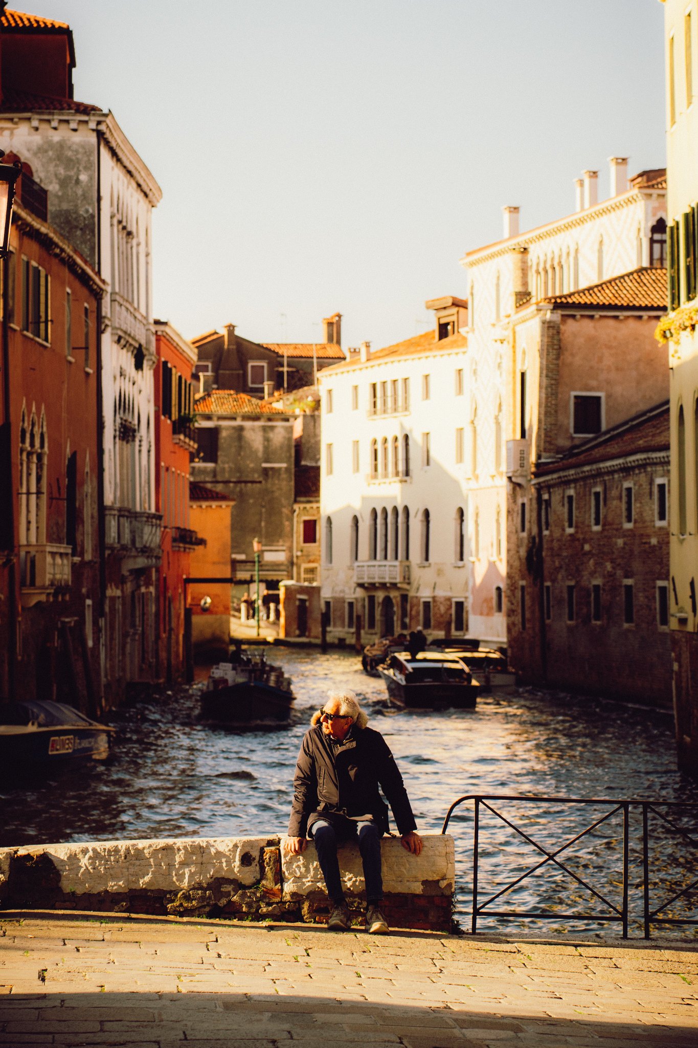 Italy - An elderly person sitting on a stone ledge beside a canal in Venice, Italy, with colorful buildings and boats in the background during sunset.