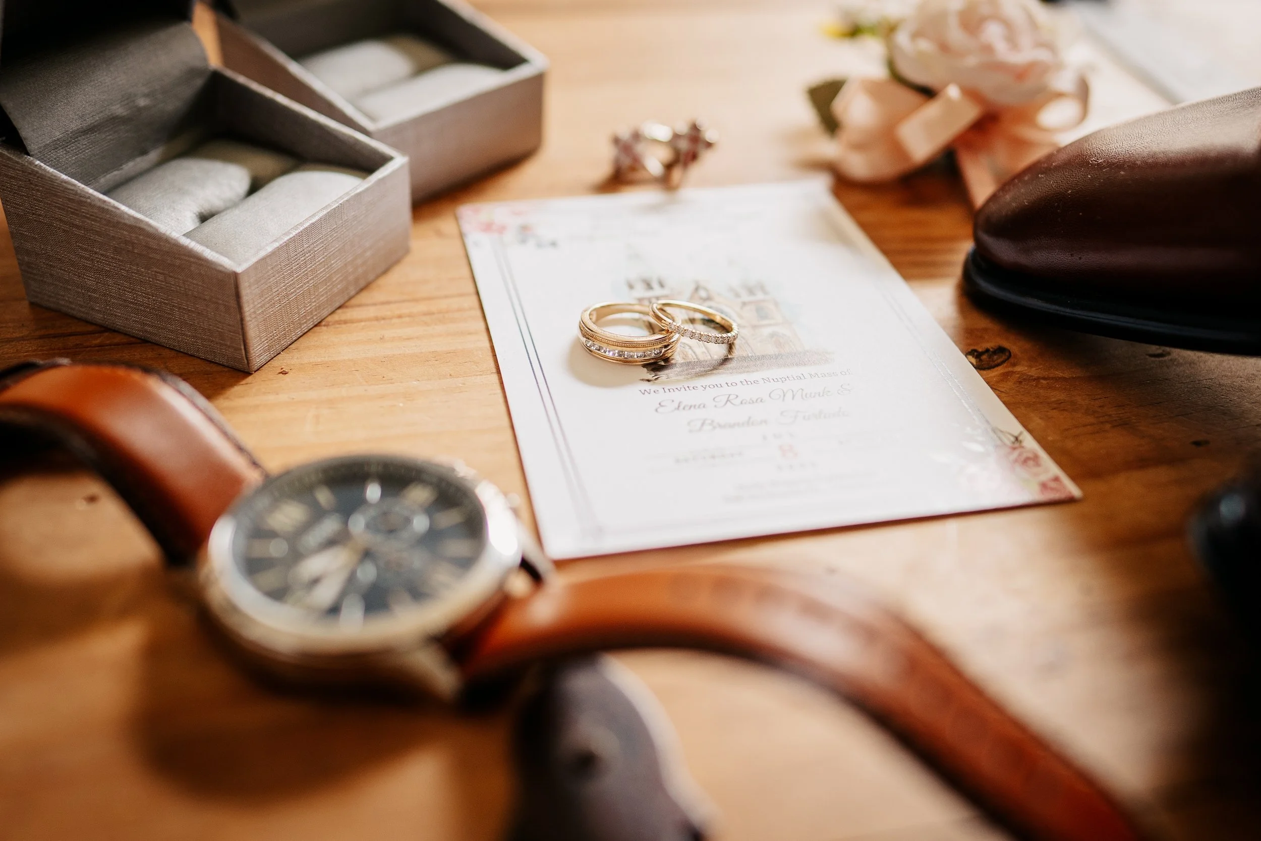 Wedding rings placed on an invitation card on a wooden surface, with watch, jewelry boxes, and accessories surrounding them.