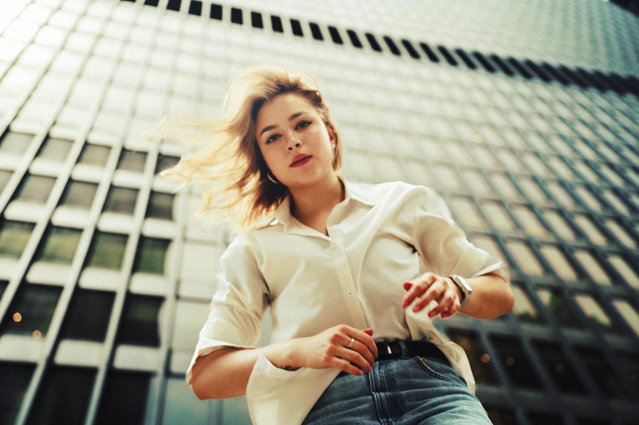 A young woman with blonde hair, red lipstick, and hoop earrings, wearing a white button-up shirt and blue jeans, standing in front of a tall glass office building, looking down at the camera with a confident expression.