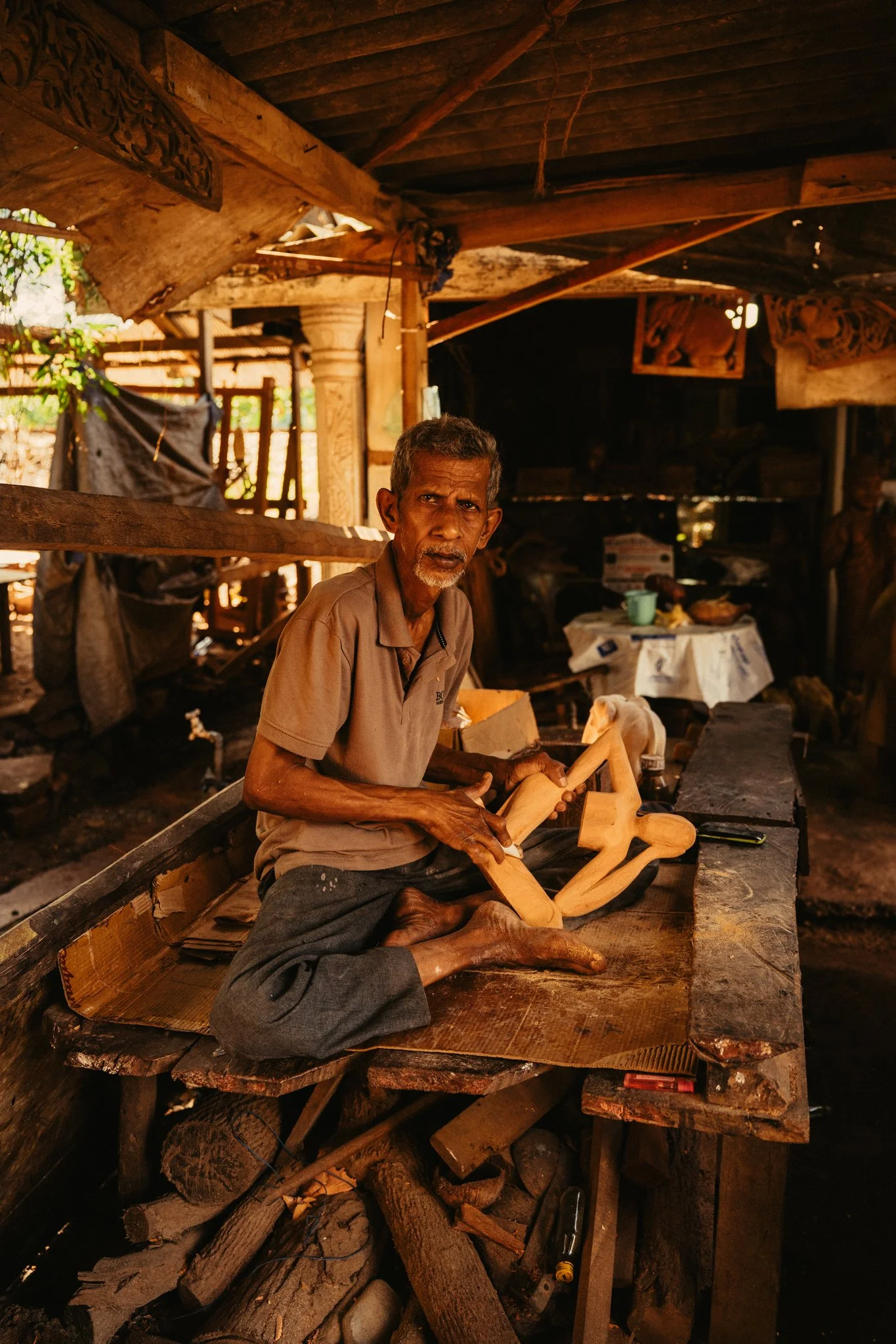 An elderly man sitting cross-legged on a wooden workbench, shaping a wooden sculpture inside a rustic workshop. The workshop has wooden beams, some carved decorative elements, and various tools and wood pieces around.