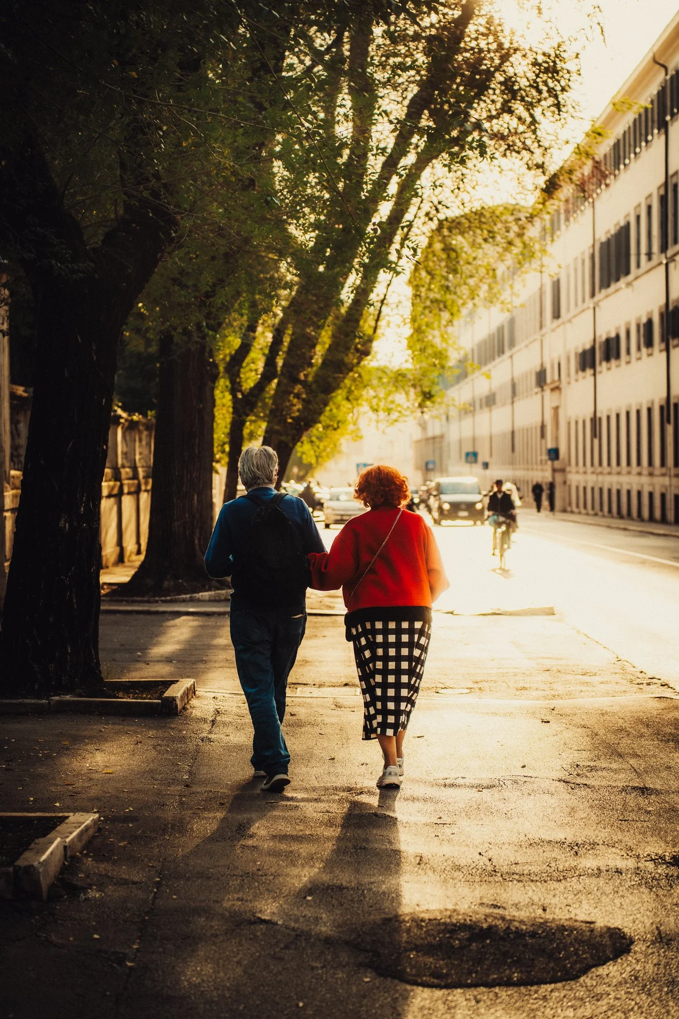Two older adults walking arm in arm down an Italian sidewalk during sunset, with trees lining the street and a large building on the right.