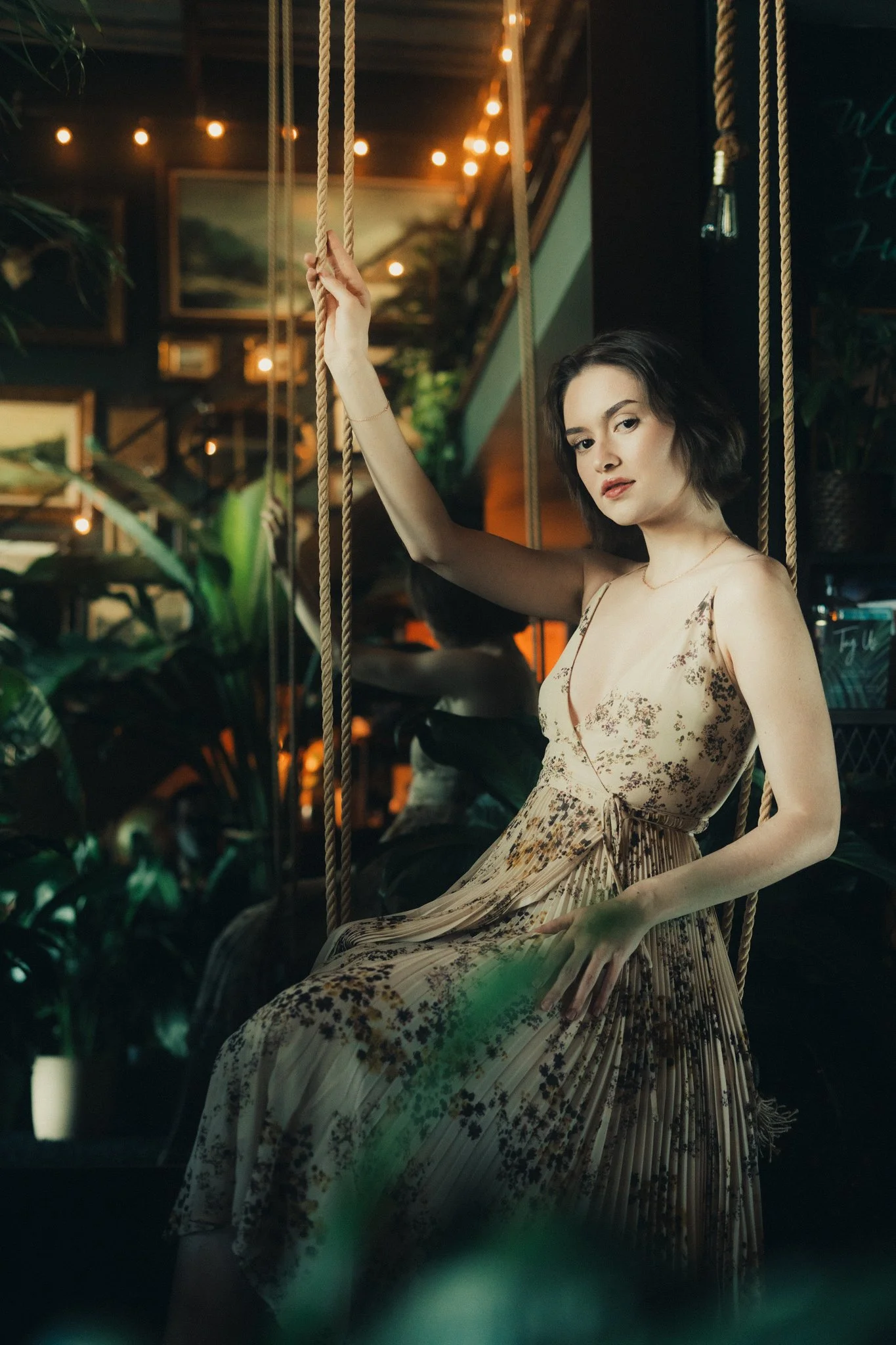 A woman in a floral dress sitting on a swing in a dimly lit indoor space with plants and warm string lights.
