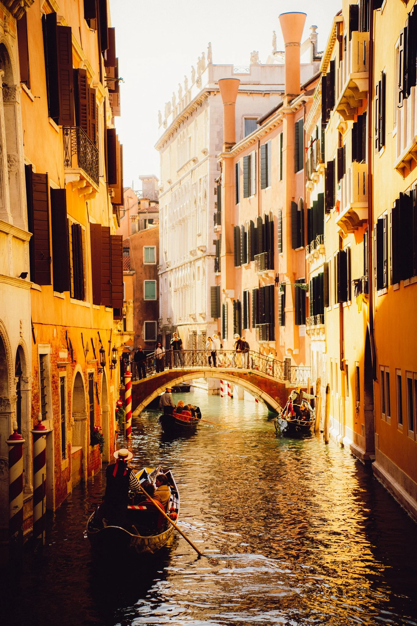 Gondolas on a narrow canal passing under a small bridge, surrounded by colorful tall buildings with black shutters, in Venice, Italy.