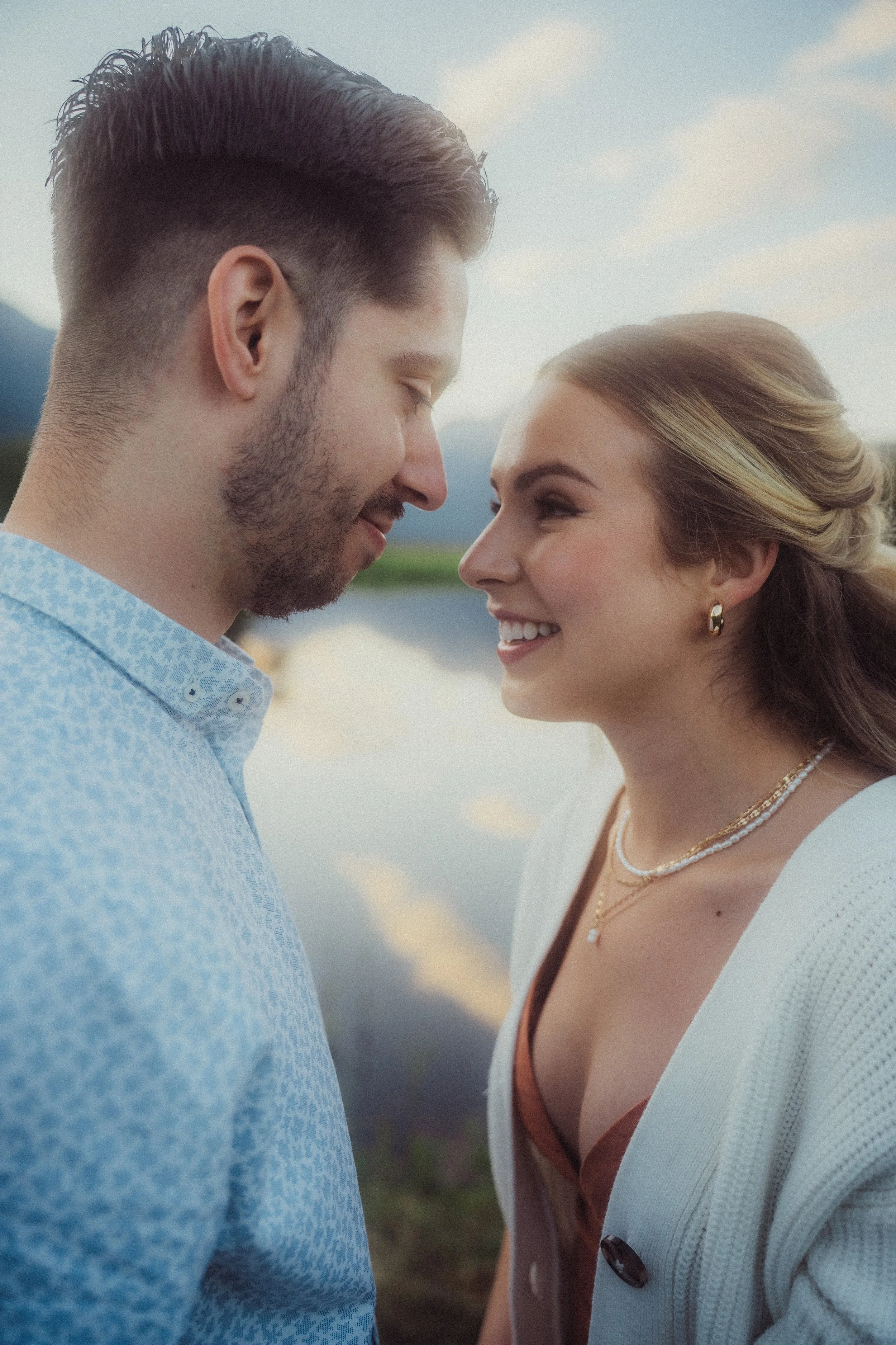 A couple standing close together outdoors near a body of water with mountains in the background, smiling at each other.