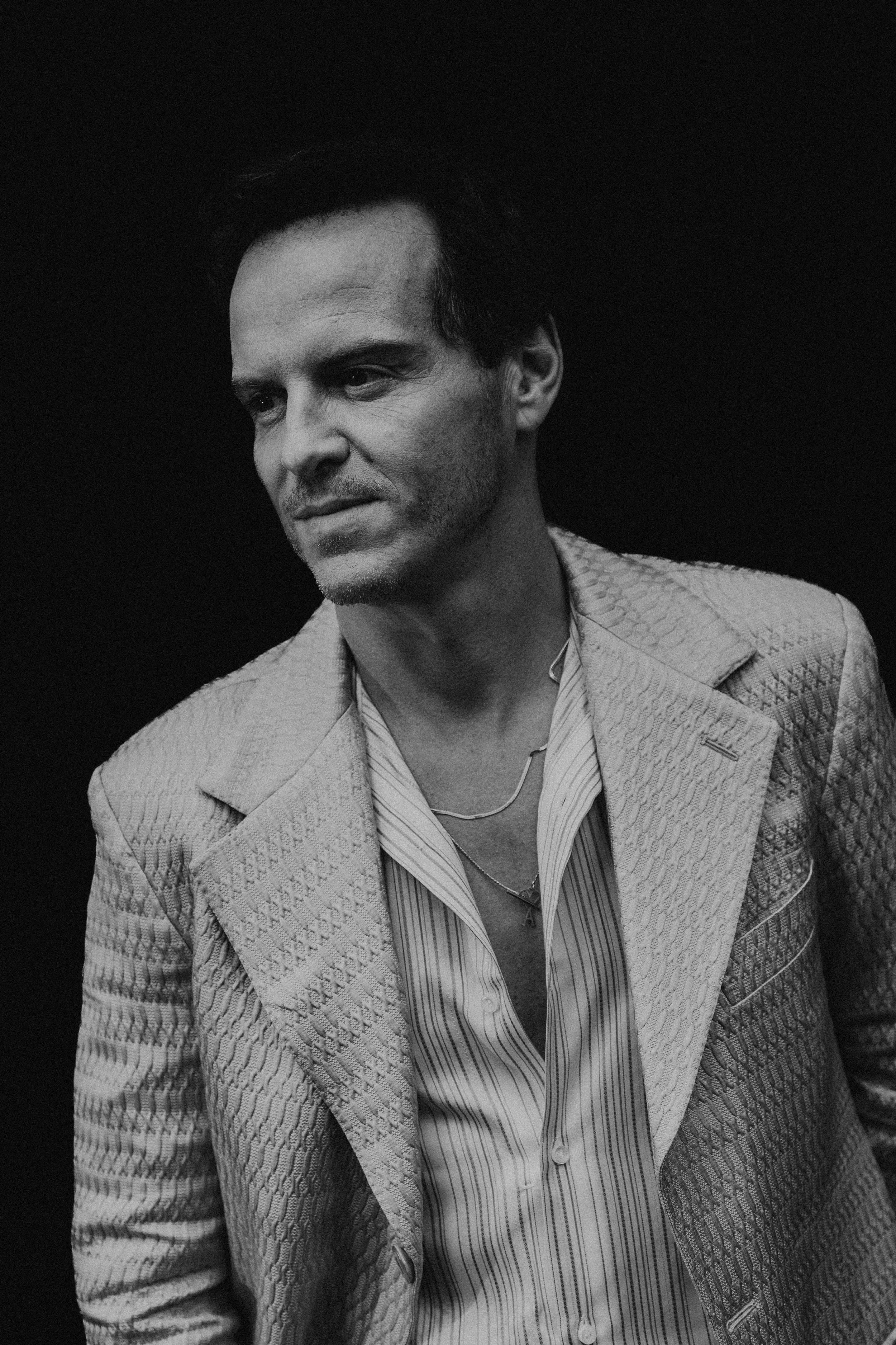 Black and white portrait of Andrew Scott looking to the side, wearing a textured blazer and a striped shirt with layered necklaces. During TIFF.