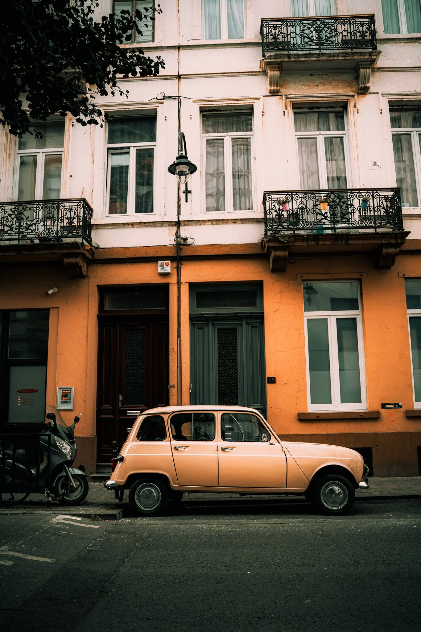 A vintage beige car parked on a city street in front of an orange building with black balconies and white windows.