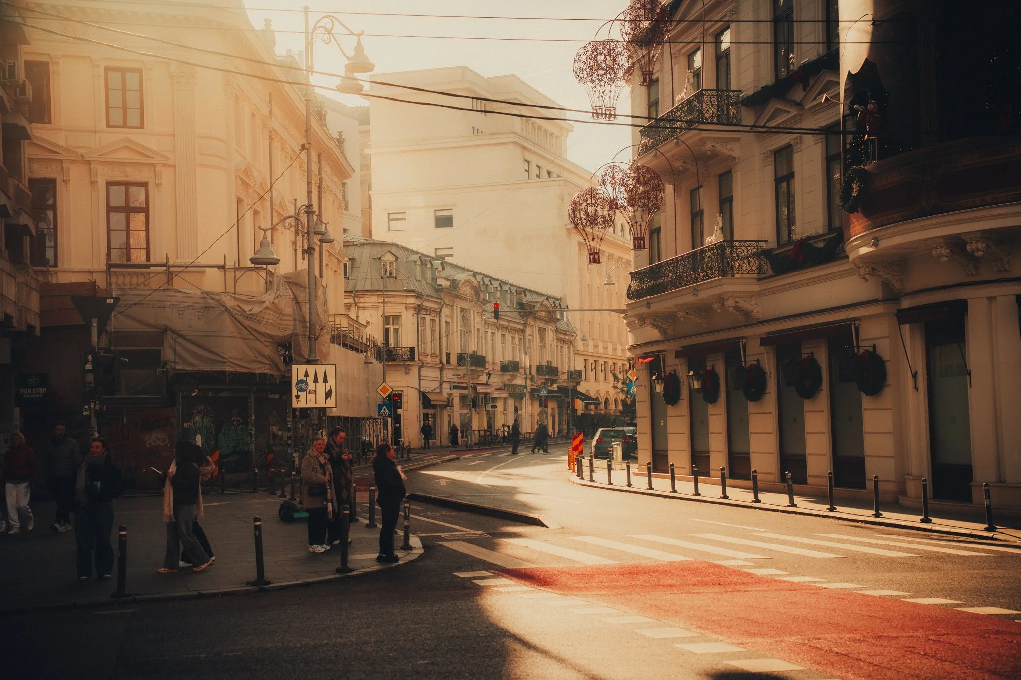 City street in the evening with people crossing at a crosswalk, classic European-style buildings, hanging decorative lights, and warm sunlight.