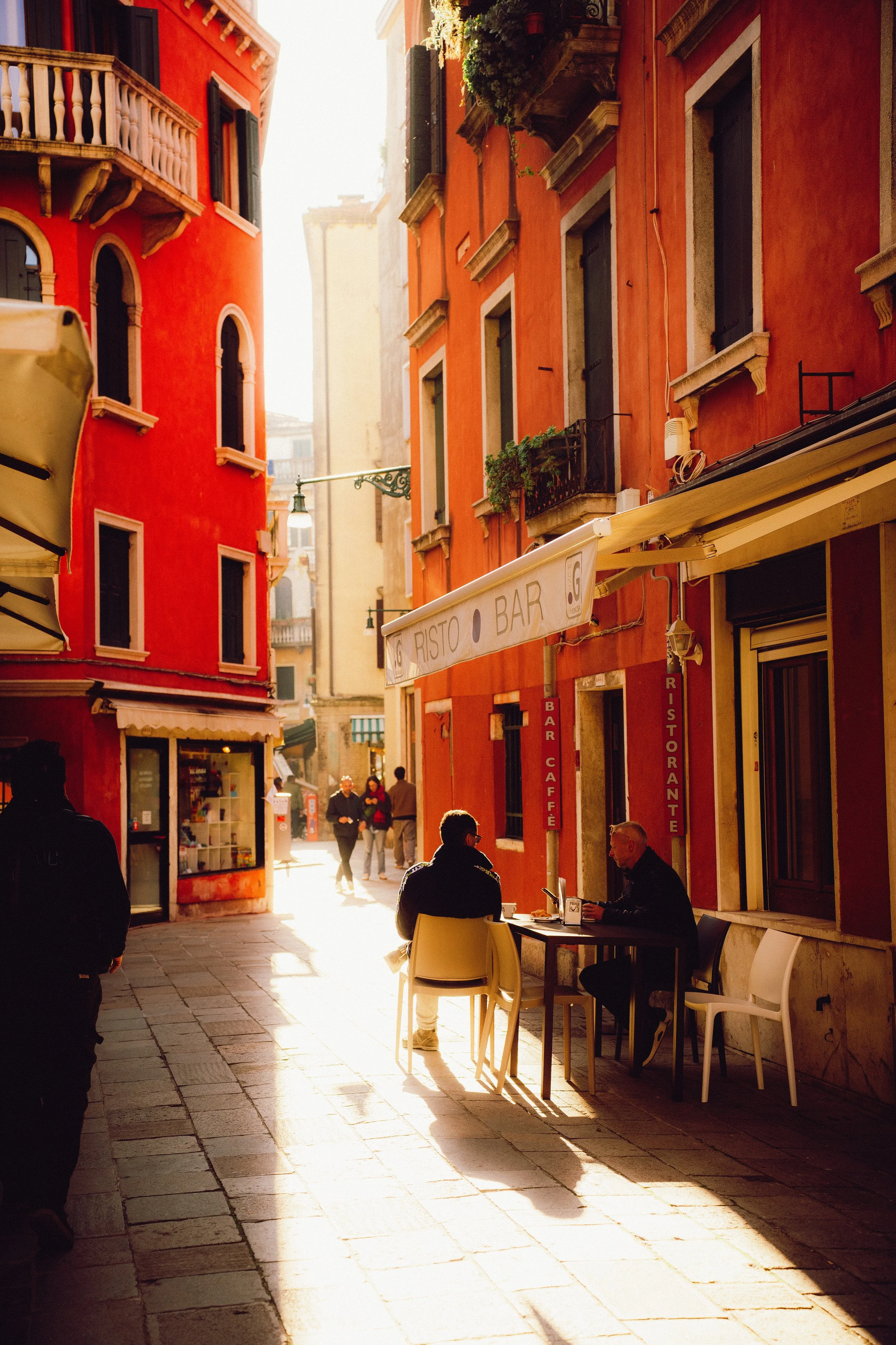 People sitting outside a cafe on a narrow European street with colorful buildings and warm sunlight.