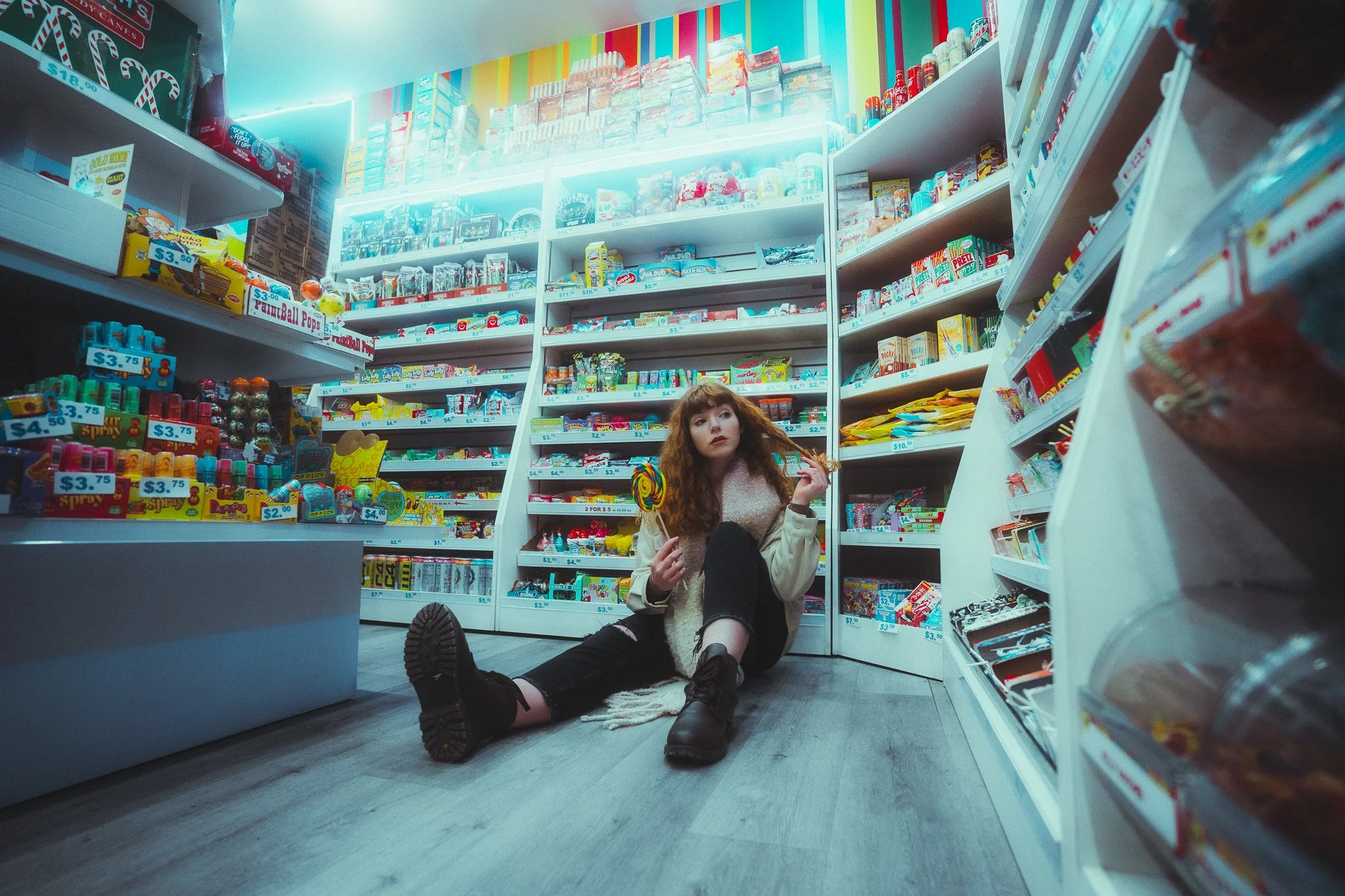 A young woman sitting on the floor of a candy store aisle, surrounded by shelves filled with various candies and snacks, holding a large lollipop in her left hand and a candy in her right hand, with a contemplative expression.