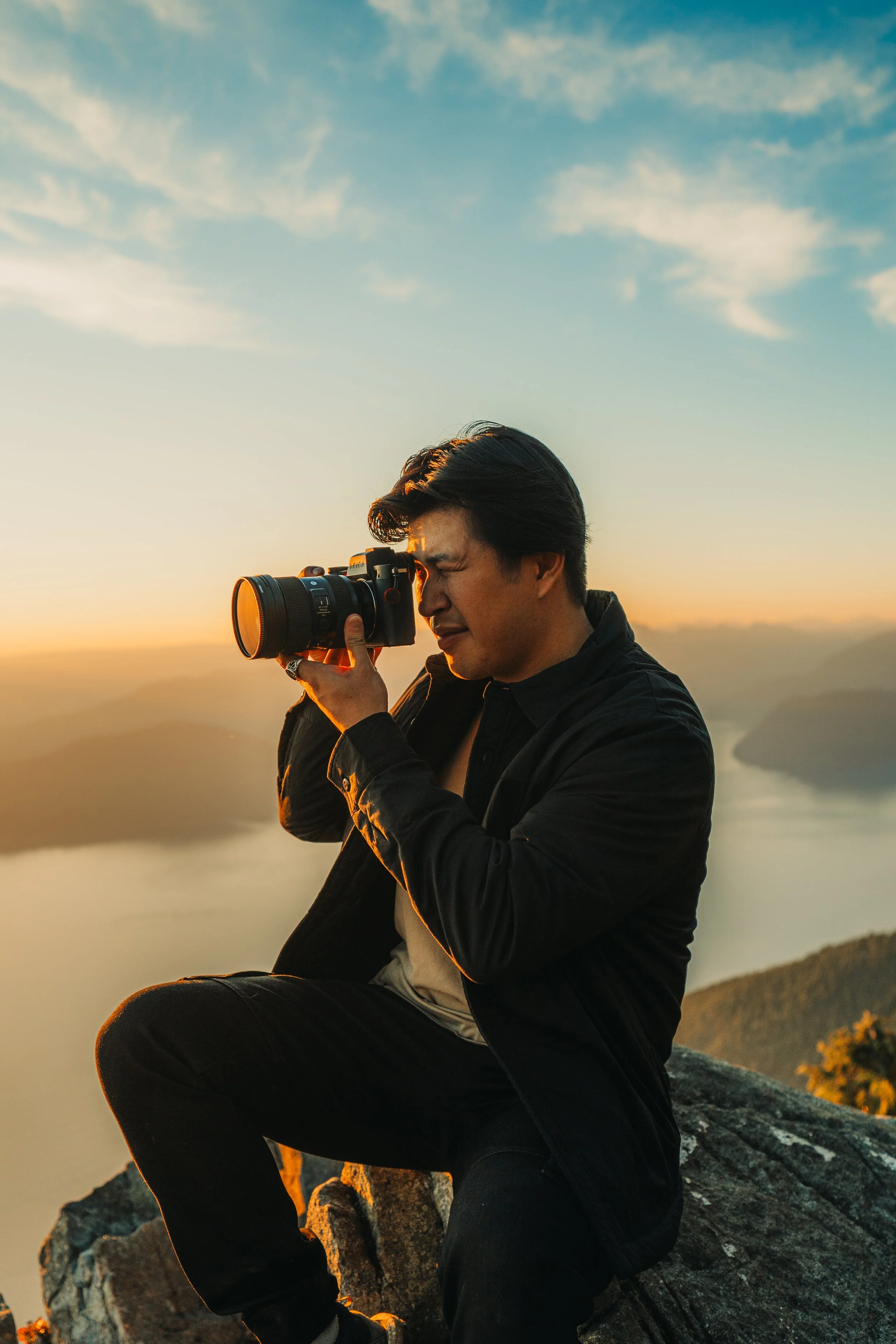 A man sitting on a rocky ledge, taking a photograph with a camera during sunset over a body of water and mountains.