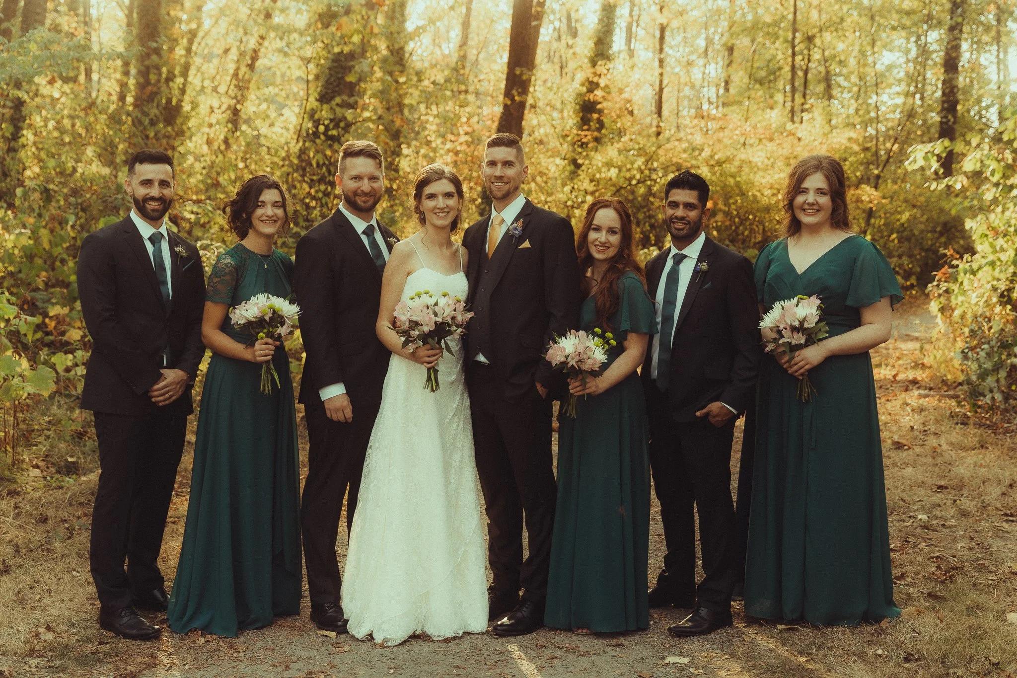 A wedding party in a forest during autumn, including a bride in a white gown, groom in a black suit with a gold tie, and six bridesmaids and groomsmen in matching dark green dresses and suits, all holding bouquets of flowers.