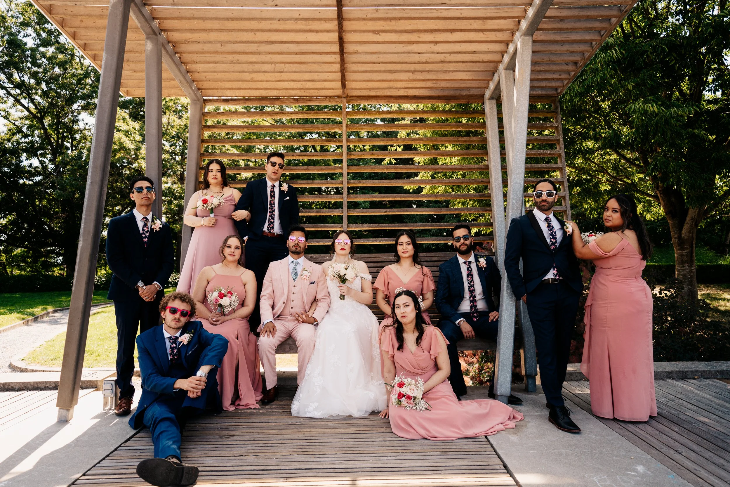 A wedding party with the bride, groom, bridesmaids in pink dresses, and groomsmen in dark suits with floral ties, posing outdoors under a wooden structure with trees in the background.