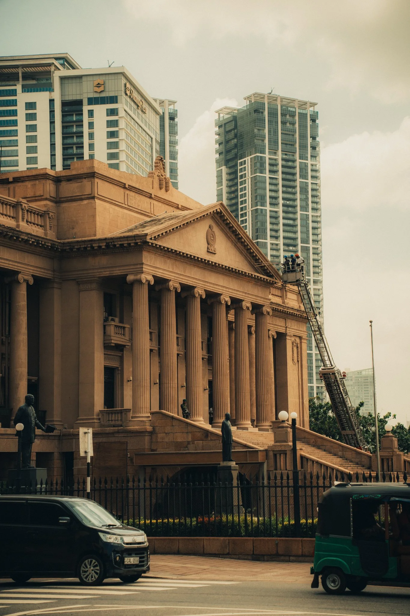 A historic government building with columns and statues in front, under construction with a crane, surrounded by modern high-rise buildings and vehicles on a street.
