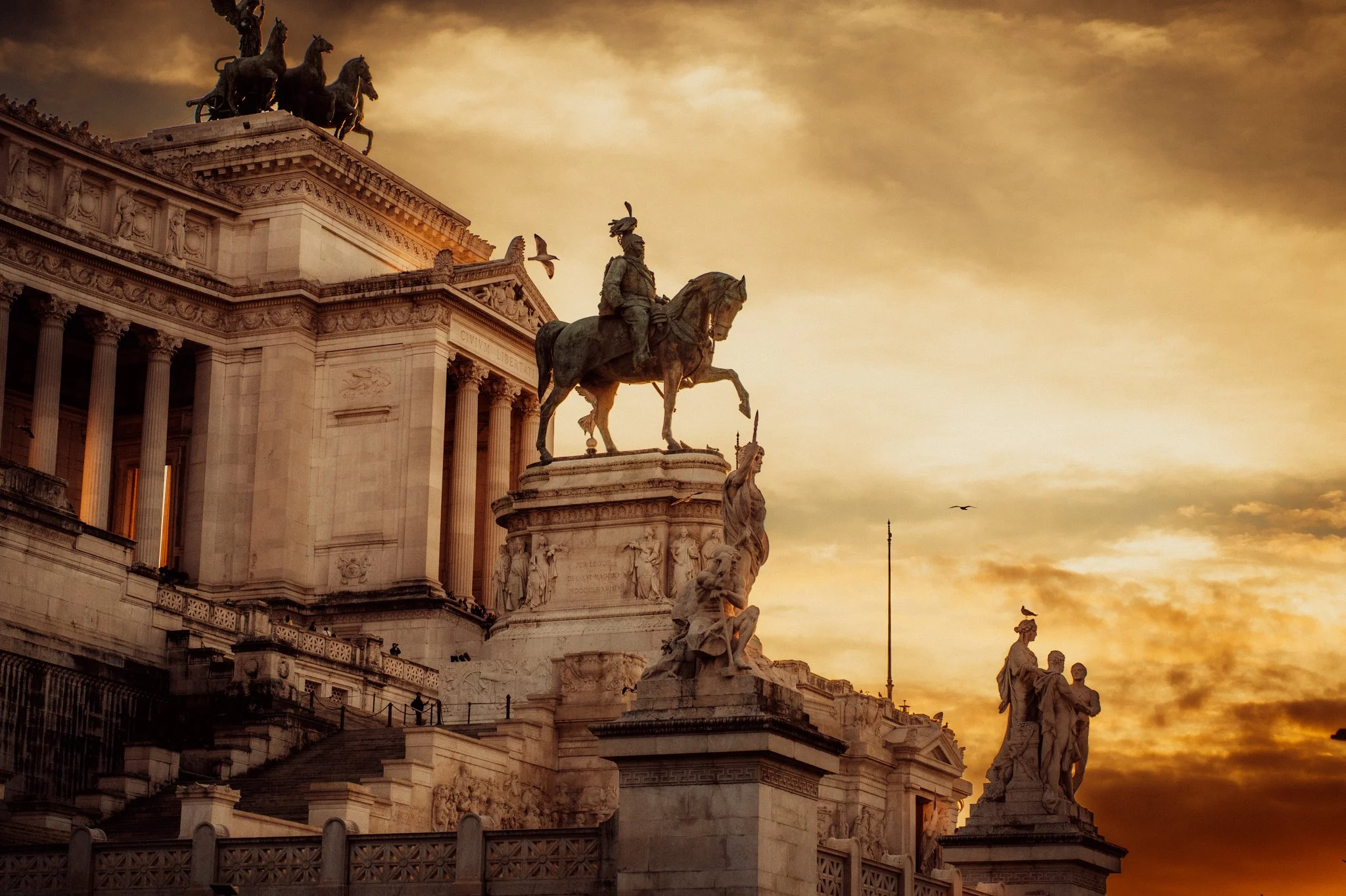 A historic monument with statues and columns during a sunset sky with clouds and birds flying.