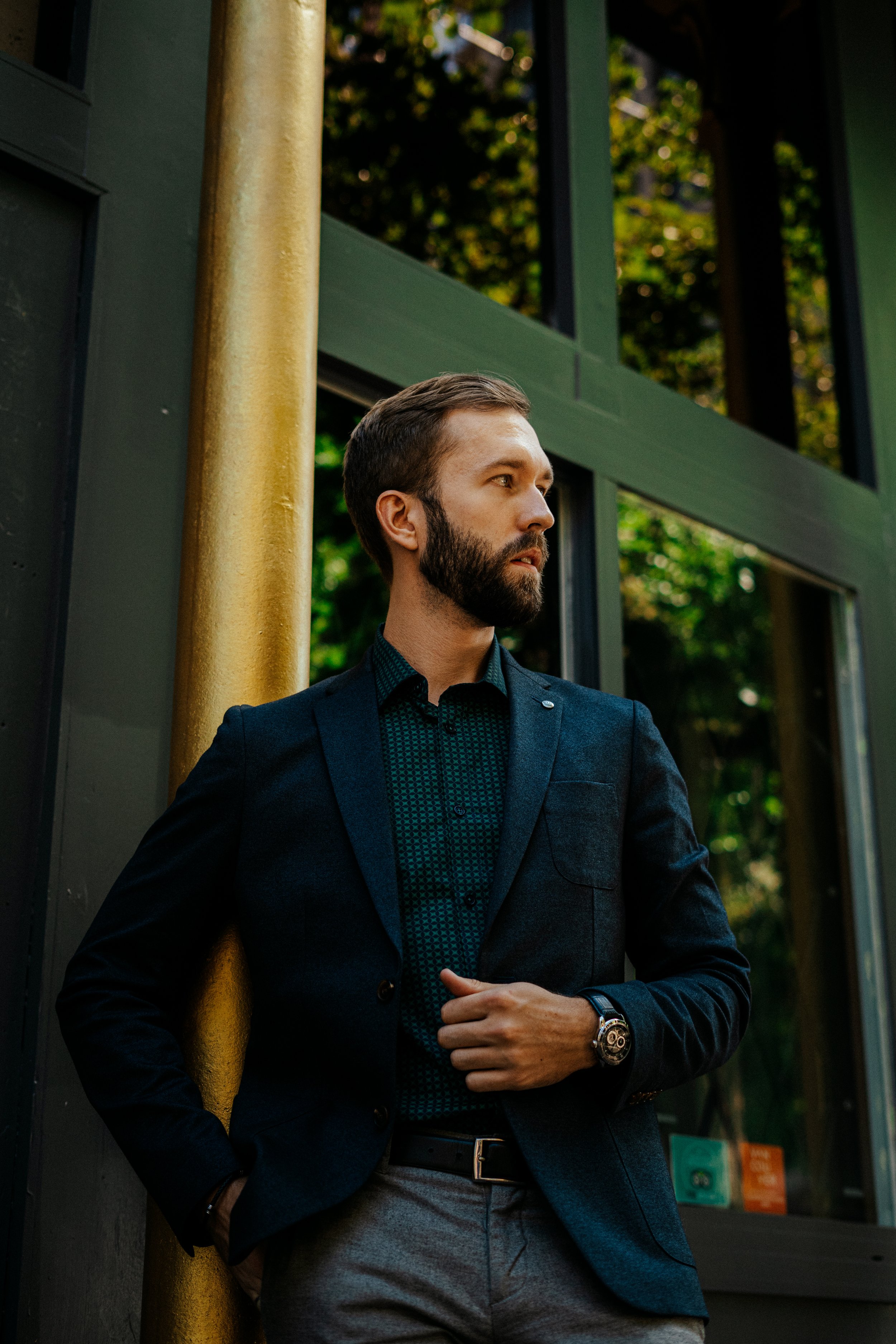 A man with a beard in a business suit standing outdoors near a green and black building, with trees in the background.