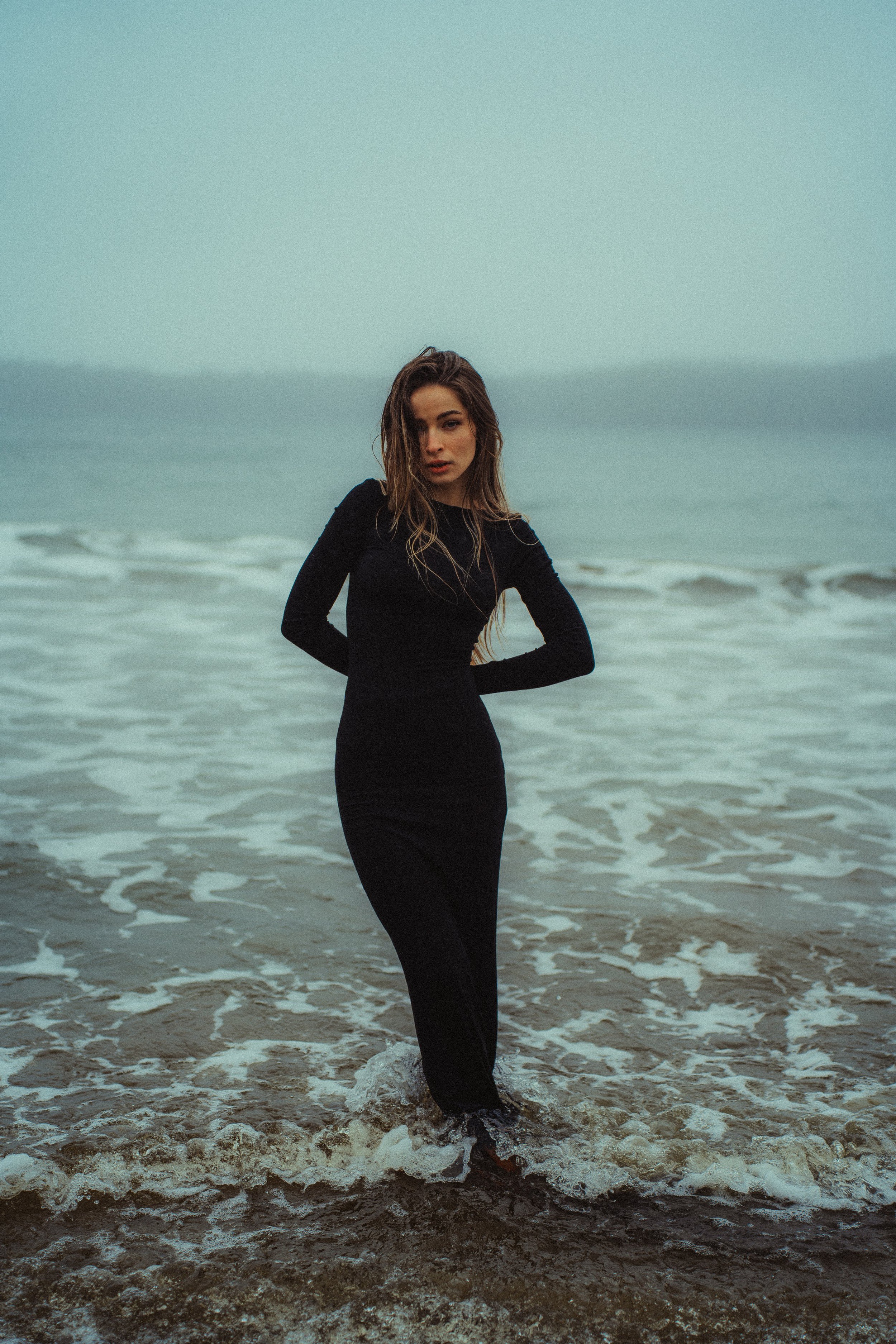 A woman wearing a black dress standing in the ocean near the shore with waves around her feet and an overcast sky in the background.