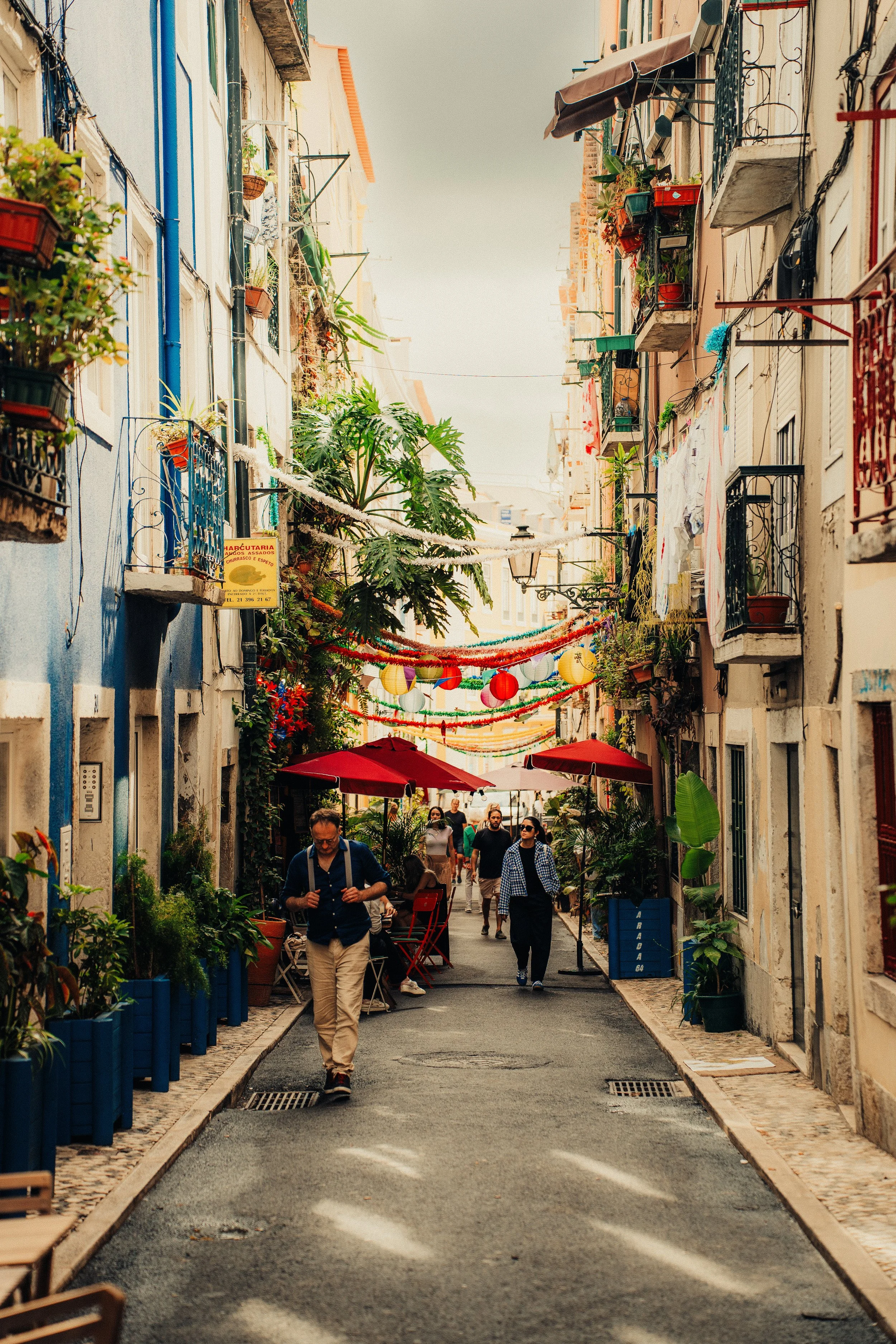 A narrow urban street decorated with colorful banners and hanging lanterns, with outdoor seating shaded by red umbrellas, and people walking and dining, lined with plants and vibrant buildings.