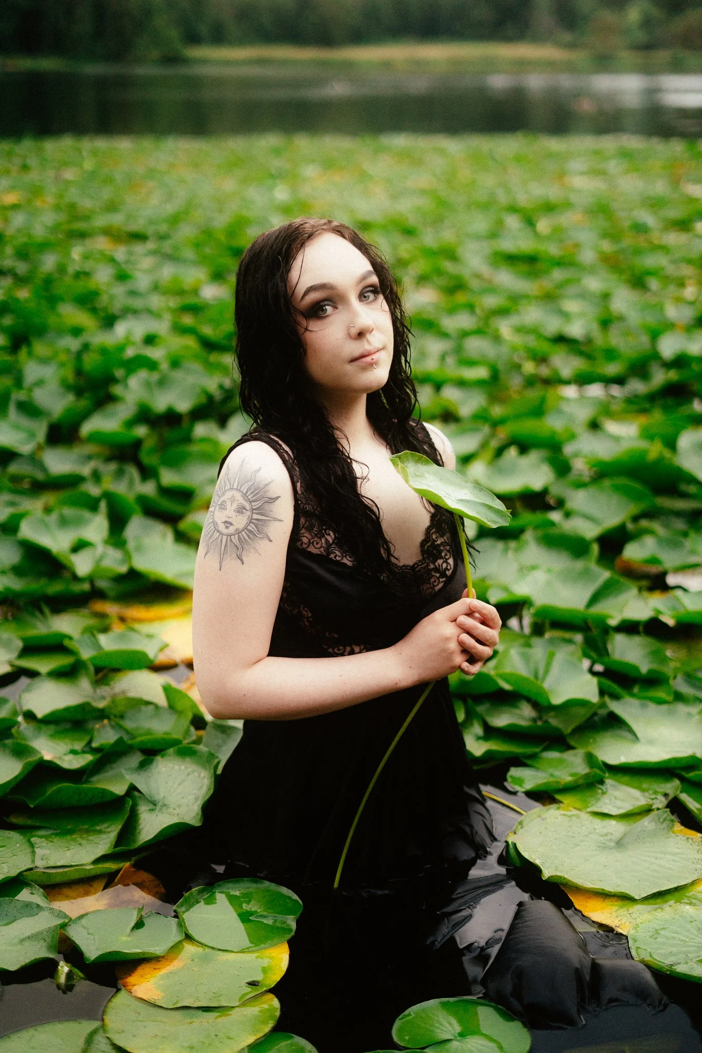 A woman with long dark hair and tattoos on her arm stands in a pond covered with green lily pads, holding a large lily pad.