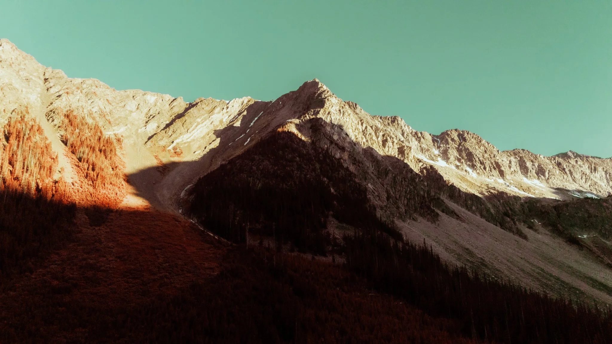 Mountain range with rocky peaks and forested slopes under a greenish sky.
