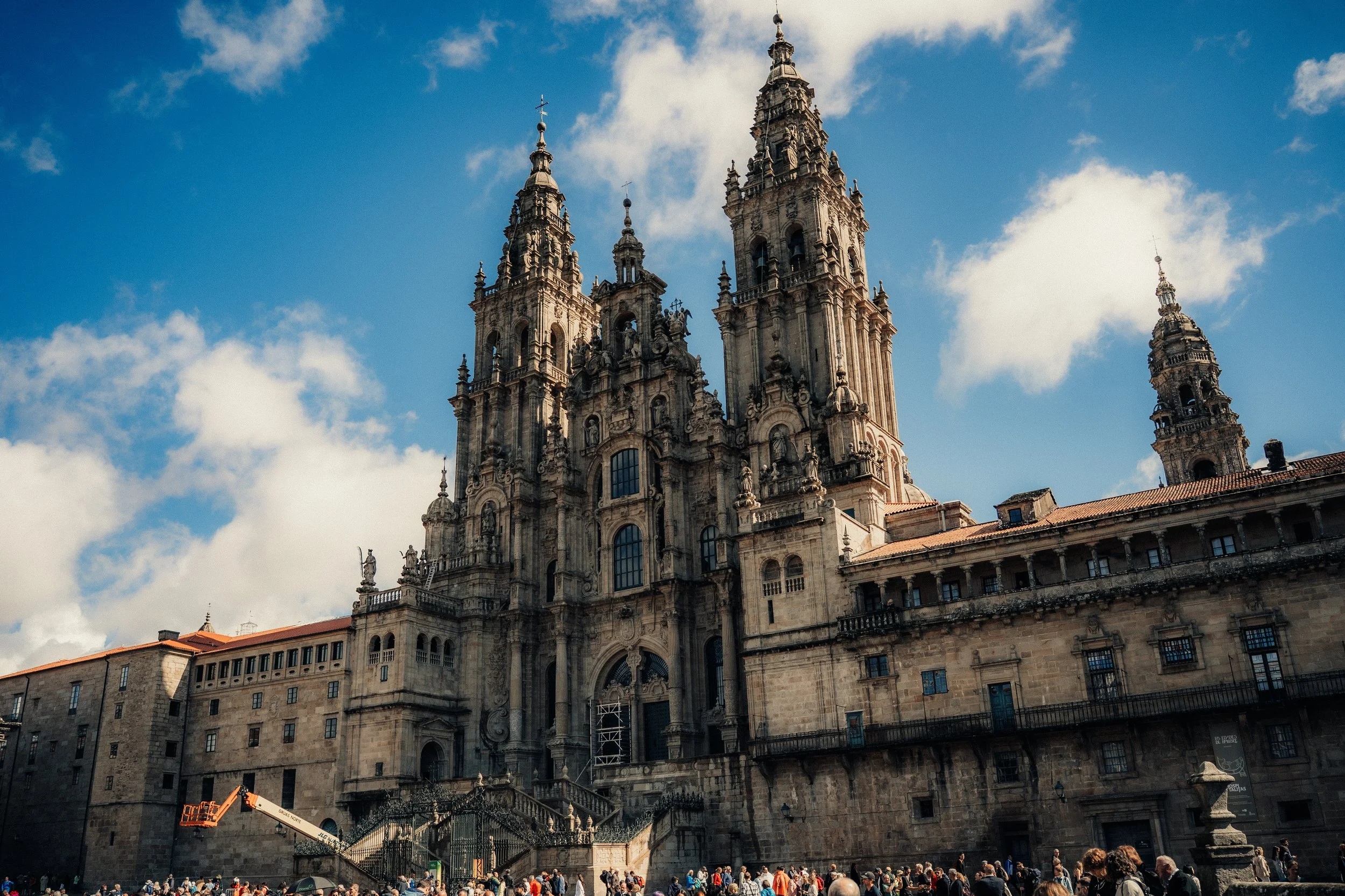 A historic cathedral with multiple tall spires and ornate architecture, set against a blue sky with clouds, with a crowd of people at the base.