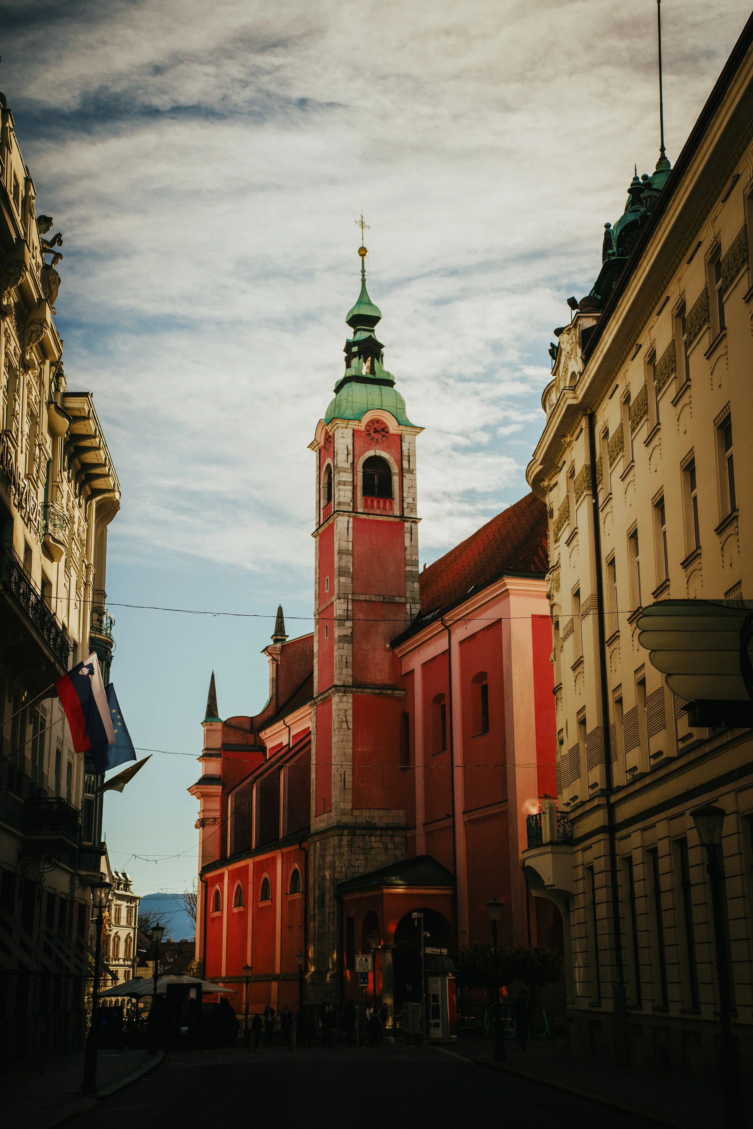 A red and pink church with a tall green spire on a city street, flanked by white buildings, with flags and street lamps in the foreground.
