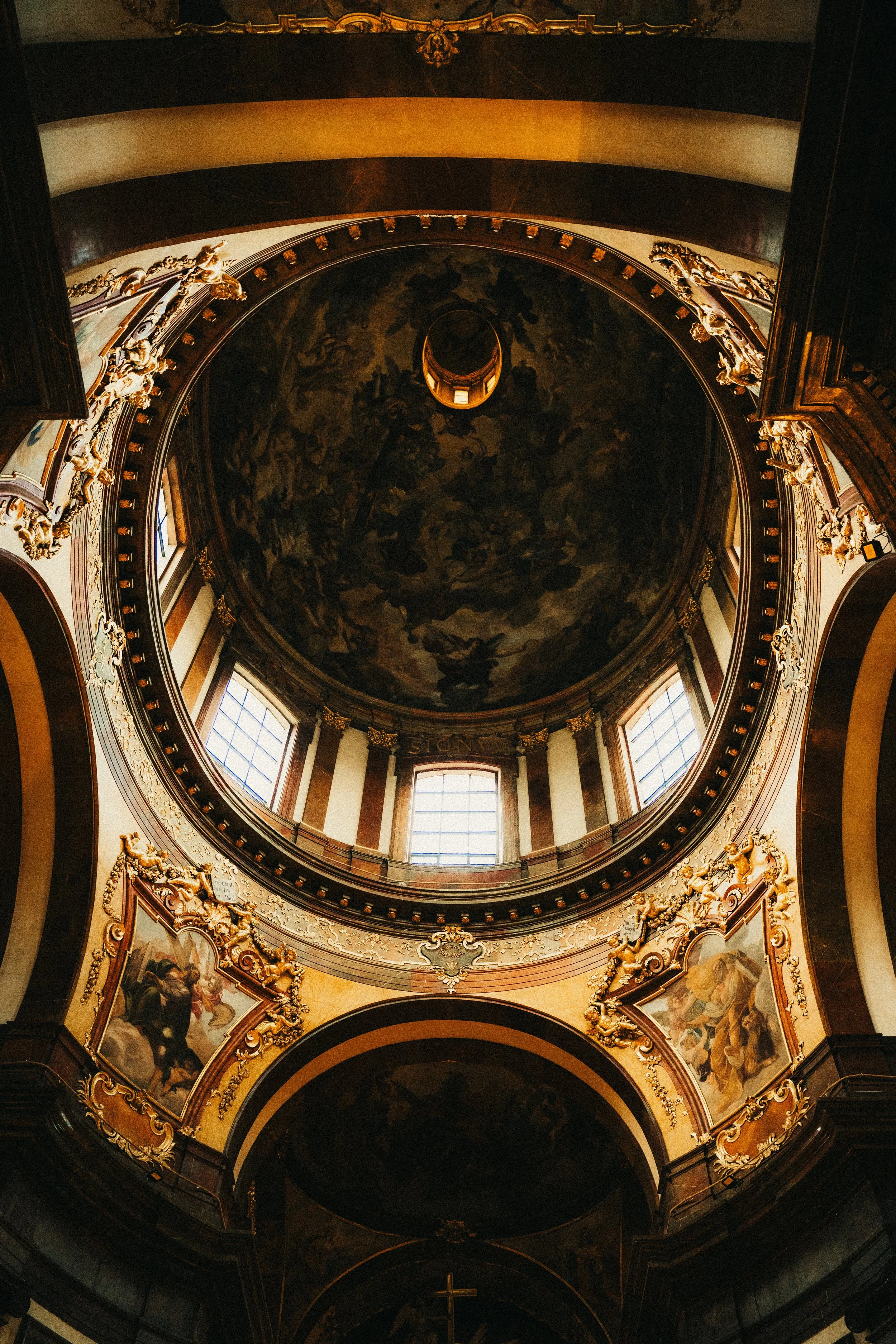 Interior view of a domed church ceiling with painted artwork, ornate gold trim, and windows letting in natural light.