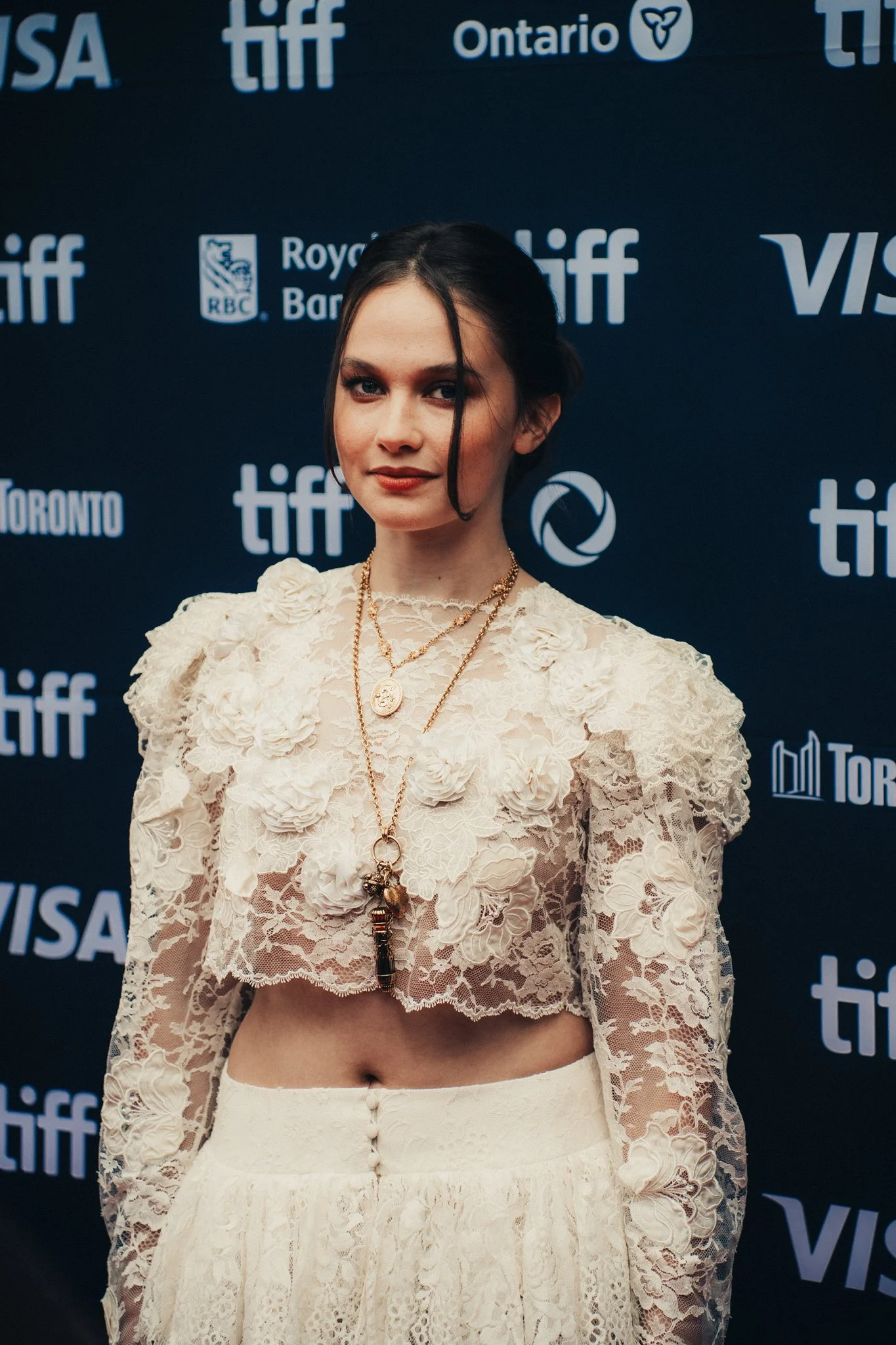 A woman in a white lace named Caliee Spaeney in a crop top and skirt posing on a blue backdrop at the Toronto International Film Festival, with various sponsor logos including TIFF, Ontario, VISA, RBC, and others.