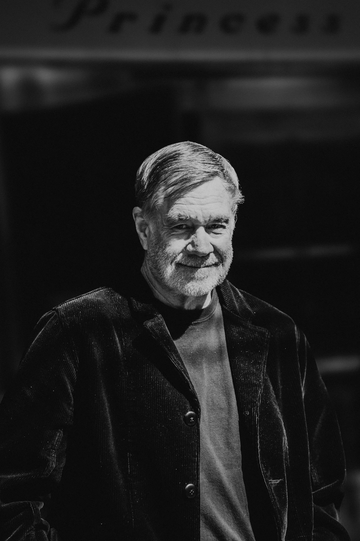 Black and white photo of Gus Van Sant wearing a dark jacket and T-shirt, smiling slightly.