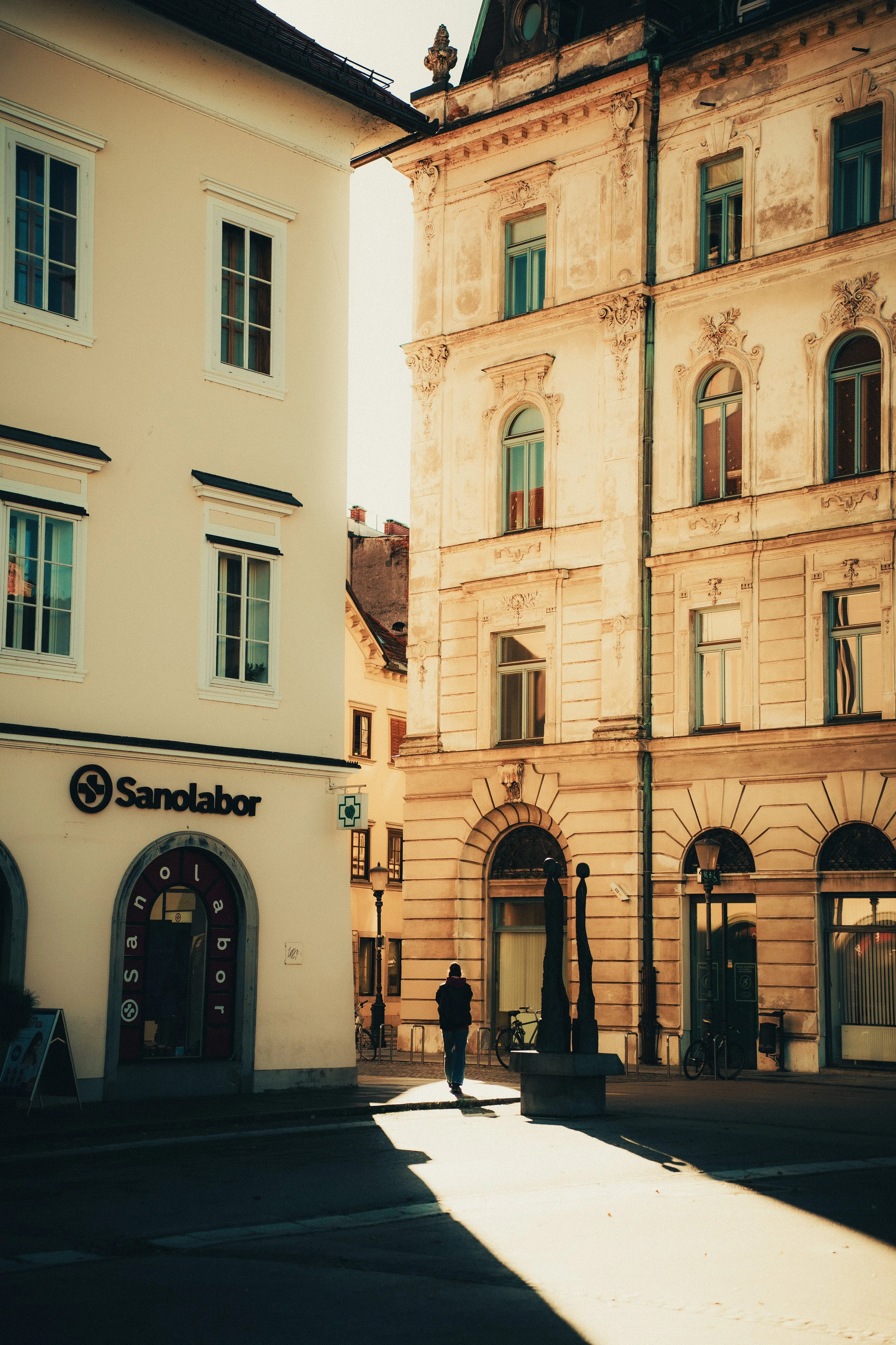 A street scene in a European city with historic buildings, including a pharmacy with a green cross sign and a salon, with one person walking and bicycles parked on the sidewalk.