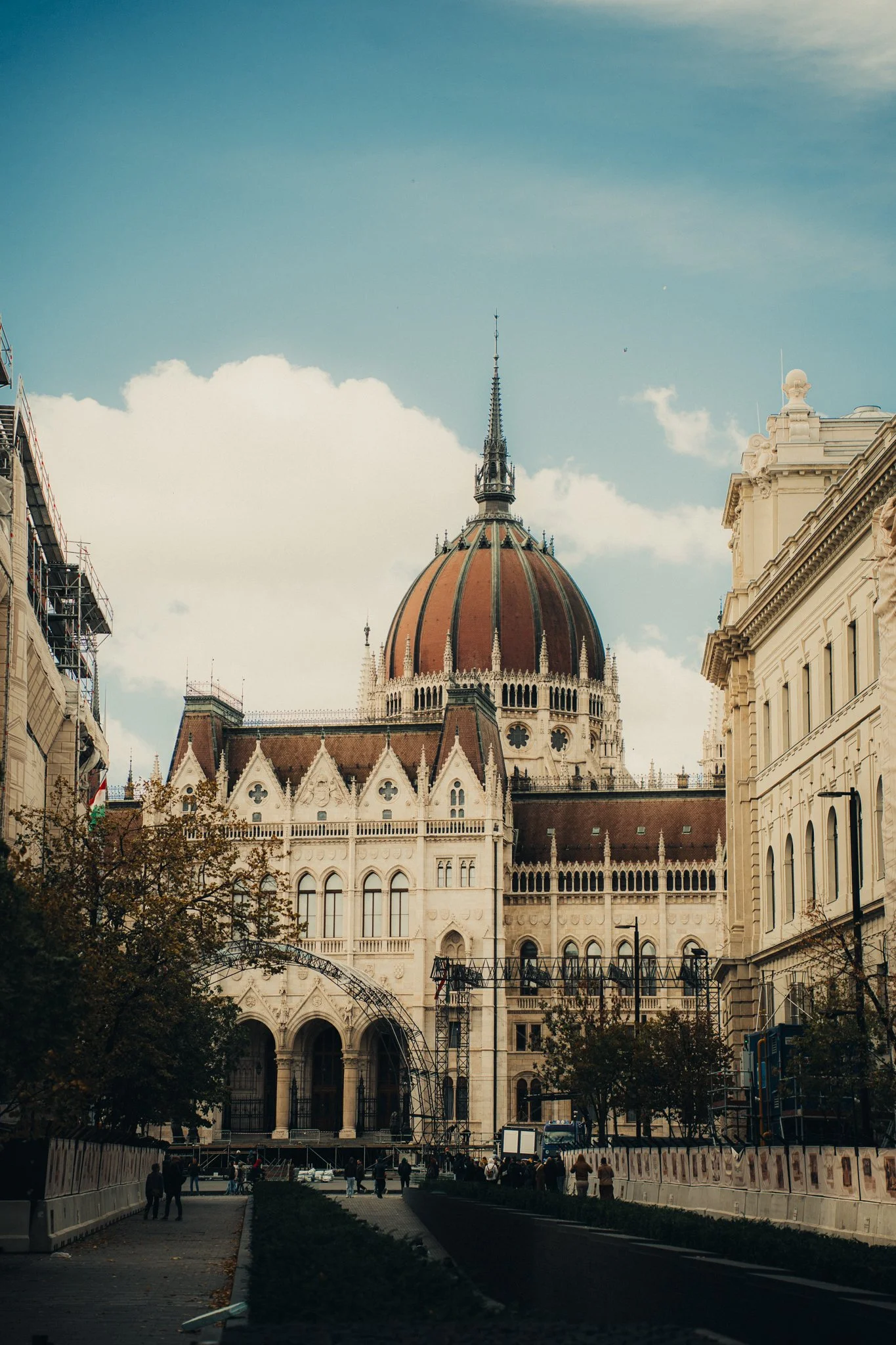 The Hungarian Parliament Building in Budapest, with its distinctive dome and Gothic Revival architecture, seen from the street with pedestrians and trees in the foreground.