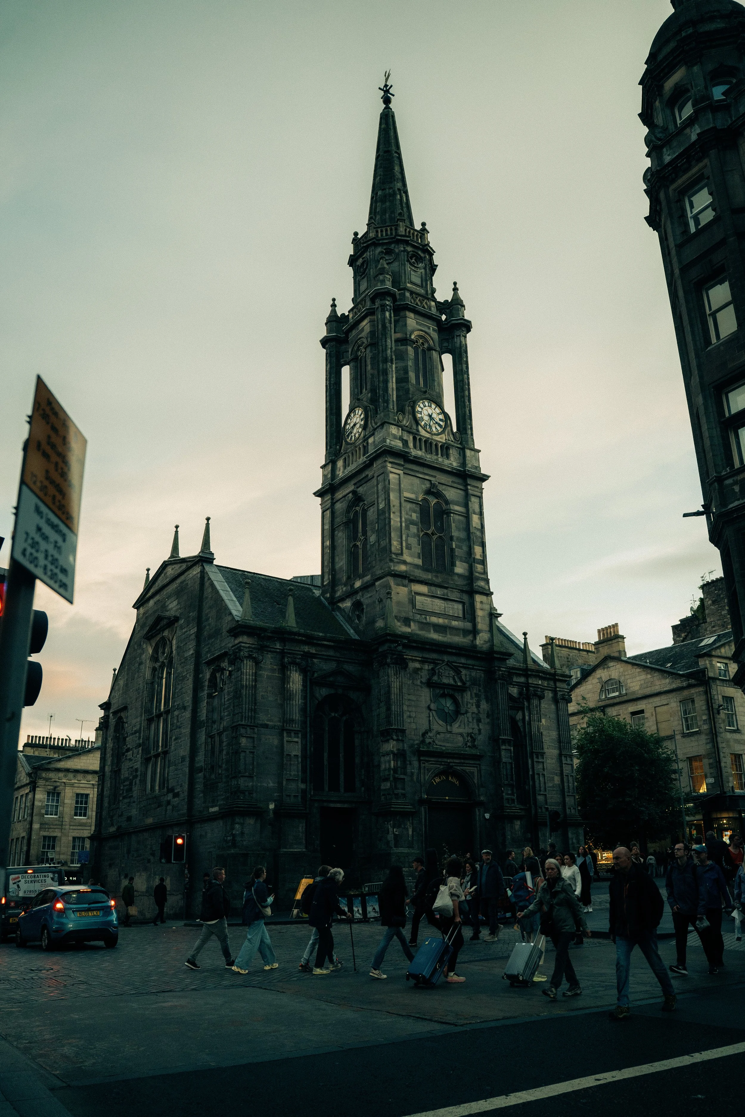 A historic church with a tall steeple and clock, surrounded by people crossing the street and carrying luggage, under an overcast sky in an urban setting.