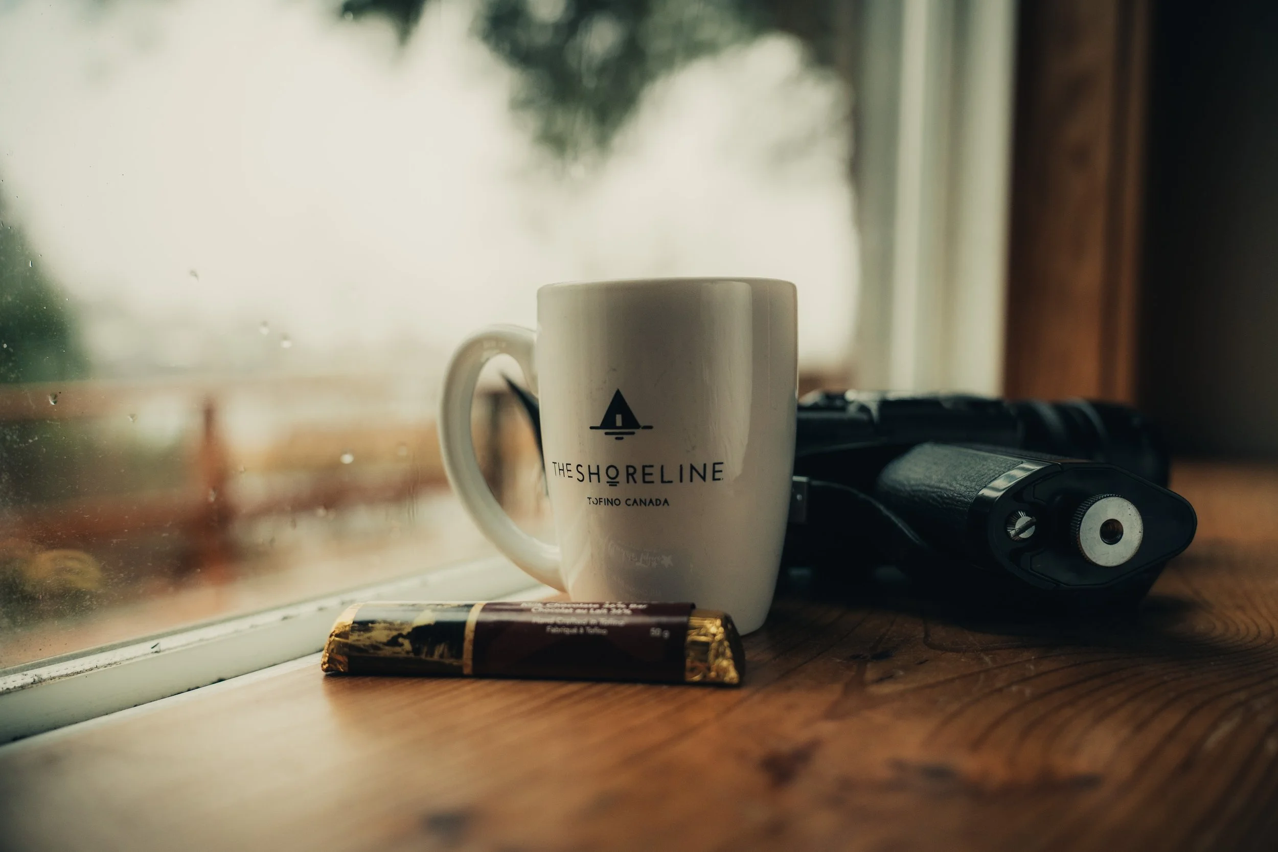 A coffee mug, a chocolate bar, and a handheld device on a wooden windowsill.