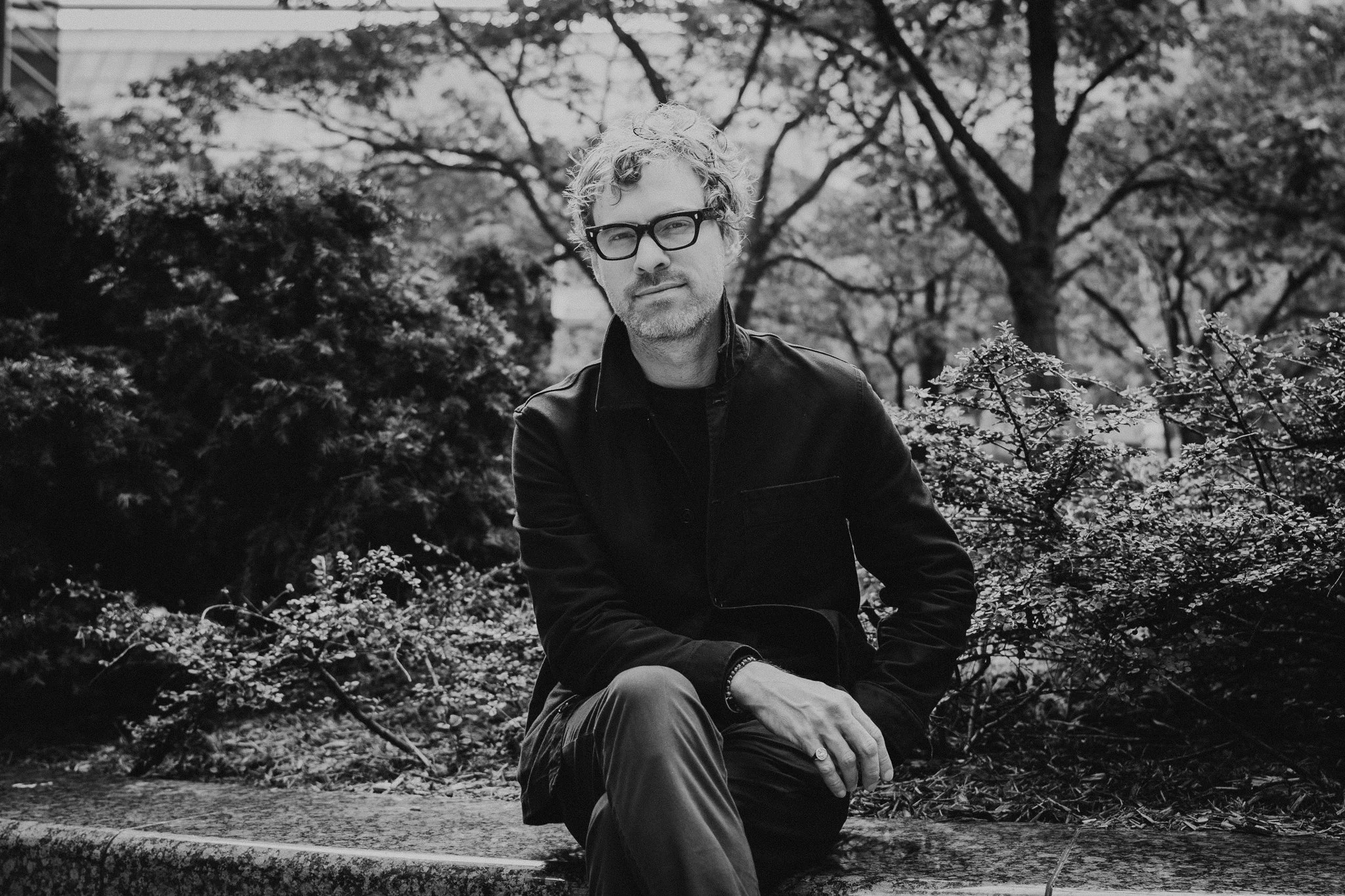 A man with glasses and curly hair is sitting on a stone ledge in a park, surrounded by trees and bushes, in black and white.