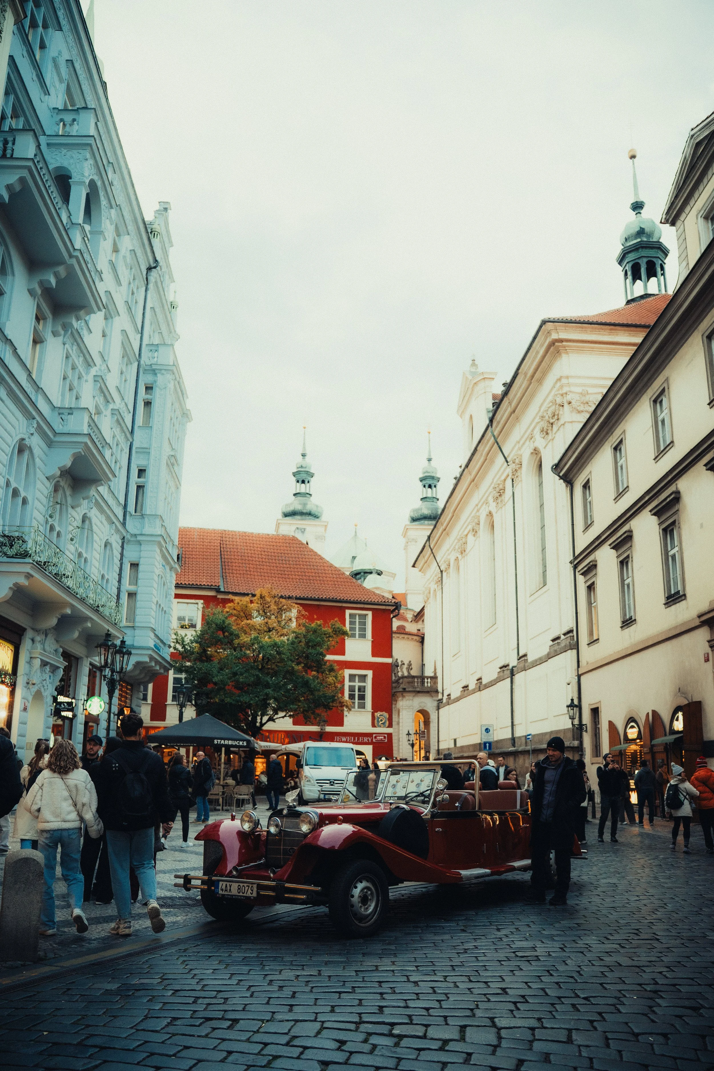 A vintage red car parked on cobblestone street in a European city square, surrounded by historic buildings and pedestrians.