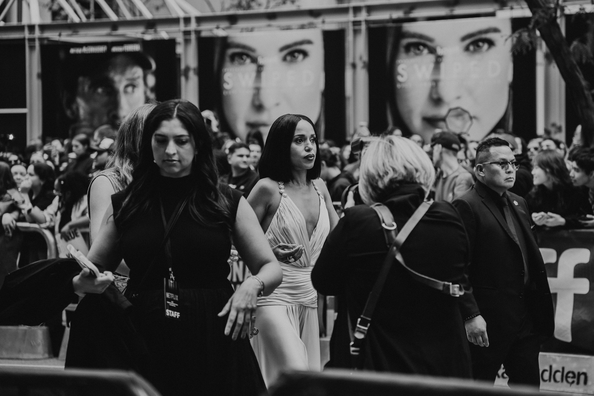 Black and white photo of a diverse group of people walking outdoors at night. Large billboard with faces and text in the background. Some individuals are wearing event badges.