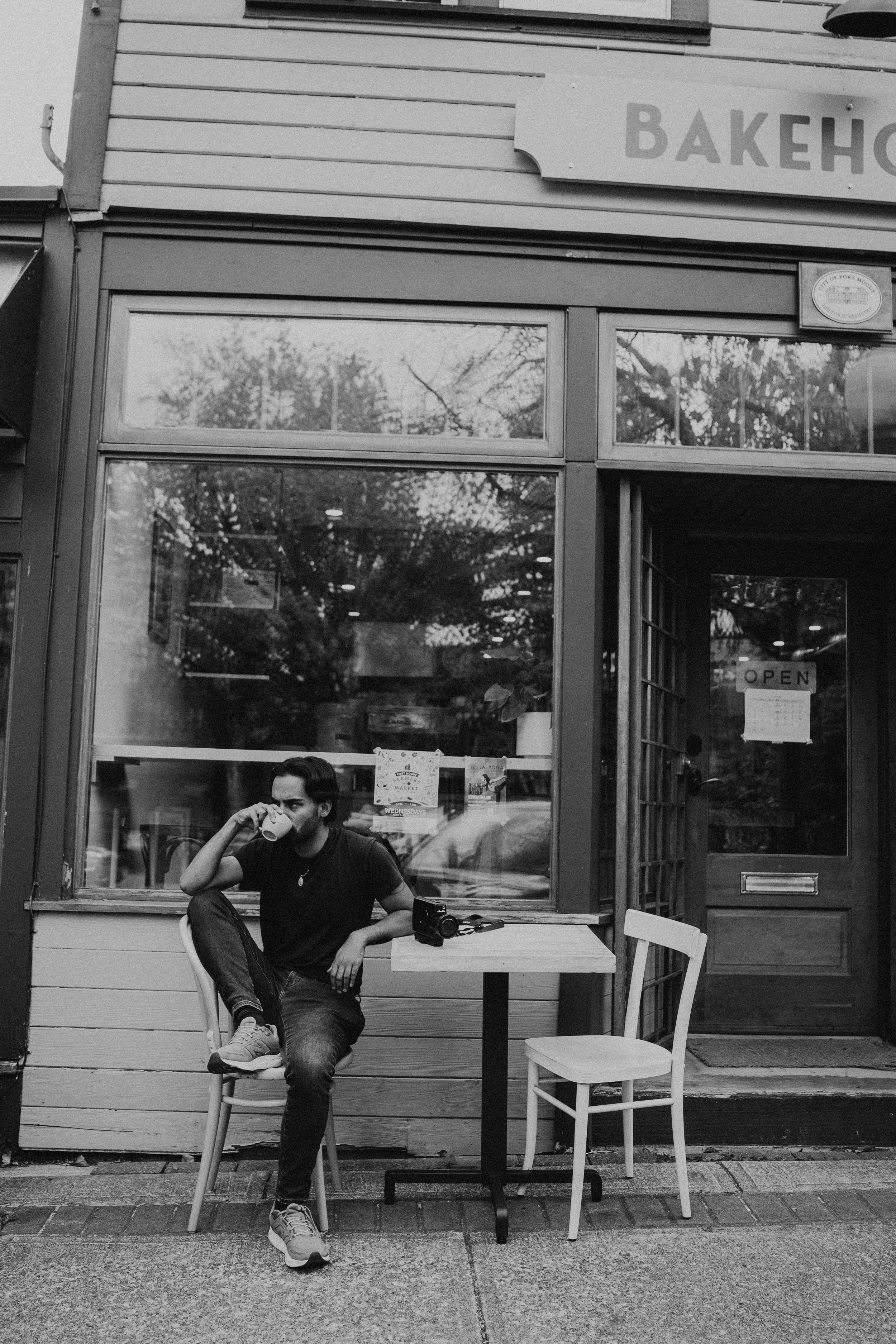 A young man with dark hair and a beard sitting outside a cafe or bakery, drinking from a mug. He is wearing a dark t-shirt, jeans, and sneakers. There is a camera on the table beside him. The building has large glass windows and a door with signs, including an "OPEN" sign. The surroundings include patio furniture and reflections of trees in the glass.