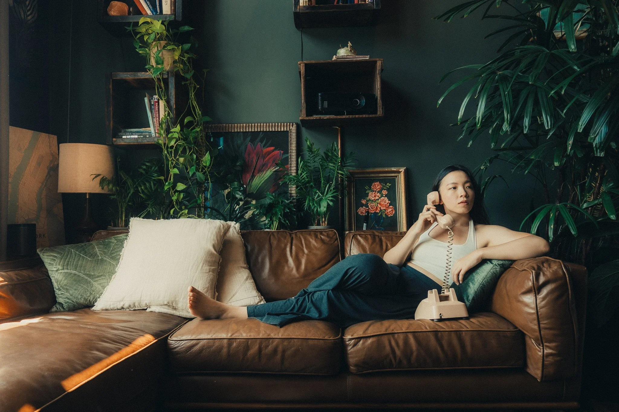 A woman lying on a brown leather sofa, talking on a beige rotary phone, in a living room with dark walls, plants, and framed art.