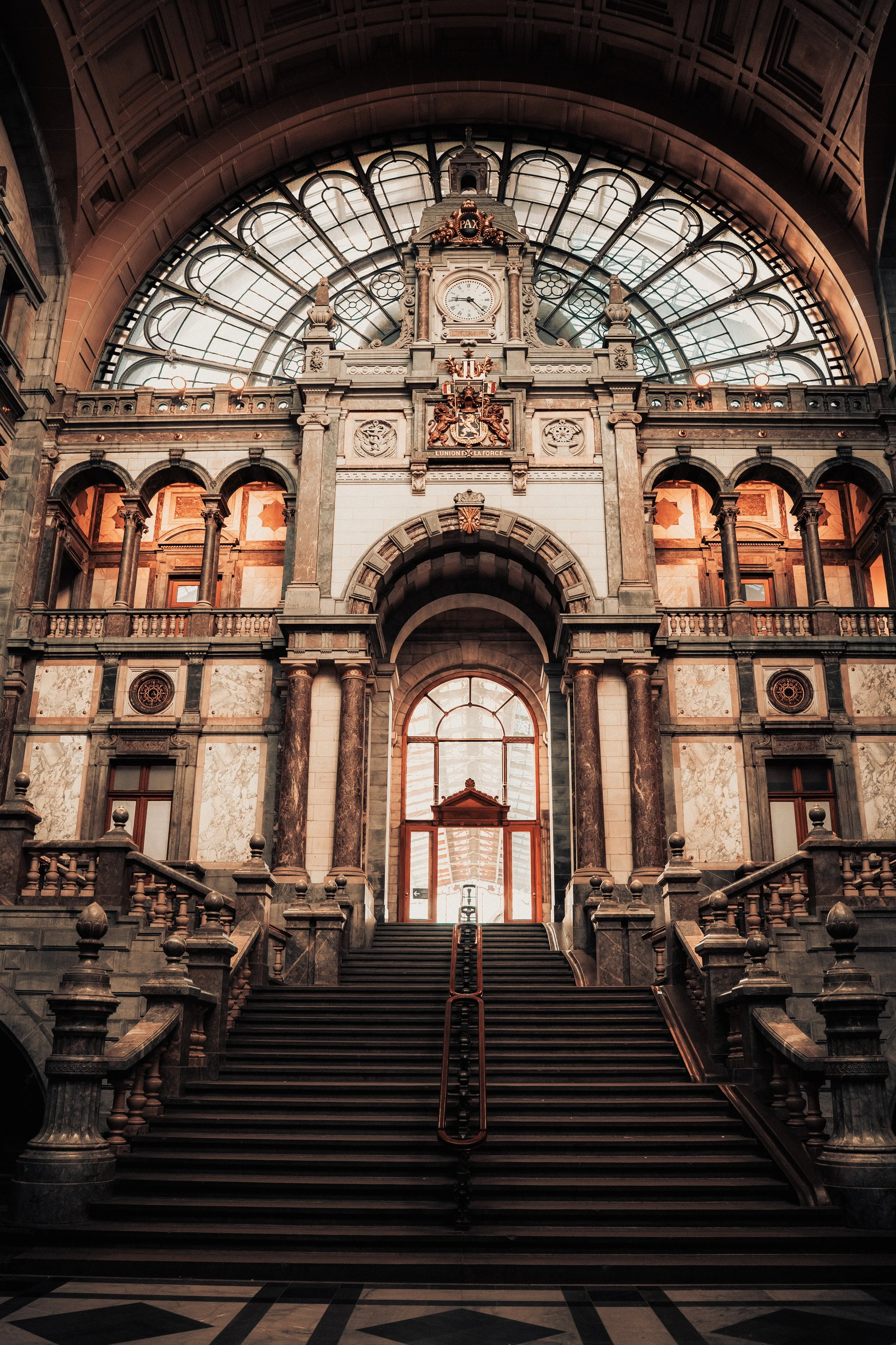 Interior view of a grand historic building with a staircase leading to large windows and an ornate clock above the archway, featuring intricate architectural details and sculptures.