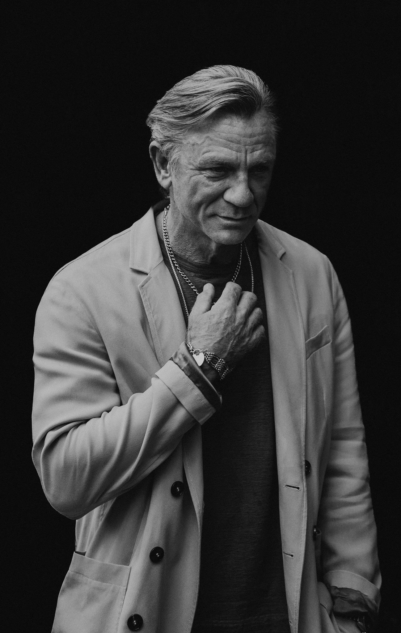 Black and white portrait of Daniel Craig with slicked-back hair, wearing a light-colored trench coat, multiple necklaces, a watch, and rings, standing against a dark background.